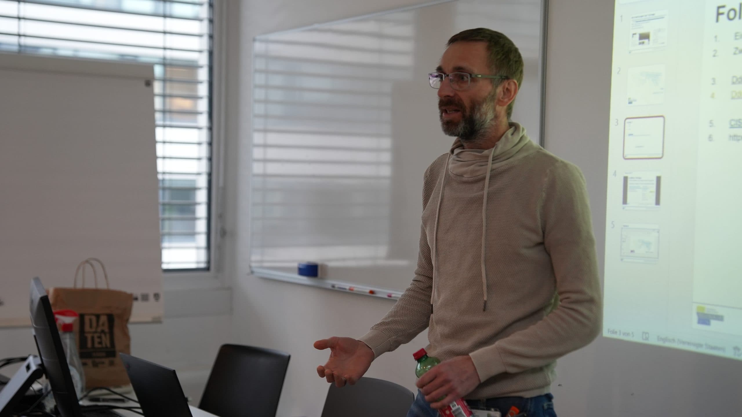 Ein Mann mit Brille und Bart spricht in einem Klassenzimmer und hält eine Flasche in der Hand. Er steht neben einer projizierten Präsentation und einem Whiteboard, auf einem Tisch sind Laptops und eine Papiertüte zu sehen.