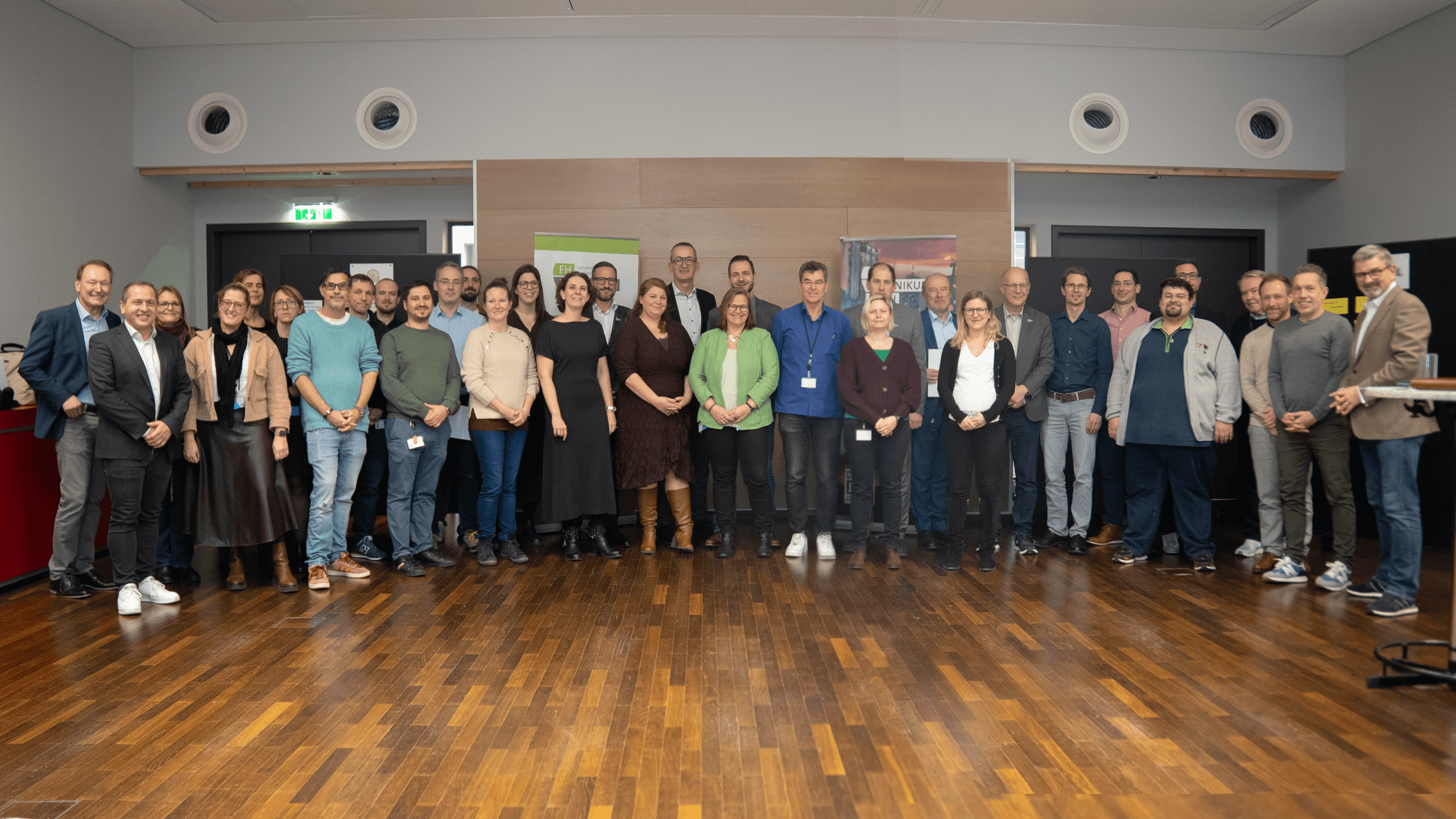 Eine große Gruppe von Menschen steht drinnen in einem Halbkreis und posiert für ein Foto. Sie sind in Freizeitkleidung gekleidet und lächeln in die Kamera. Im Hintergrund sind ein Holzfußboden und eine beigefarbene Wand mit runden Lüftungsöffnungen zu sehen.