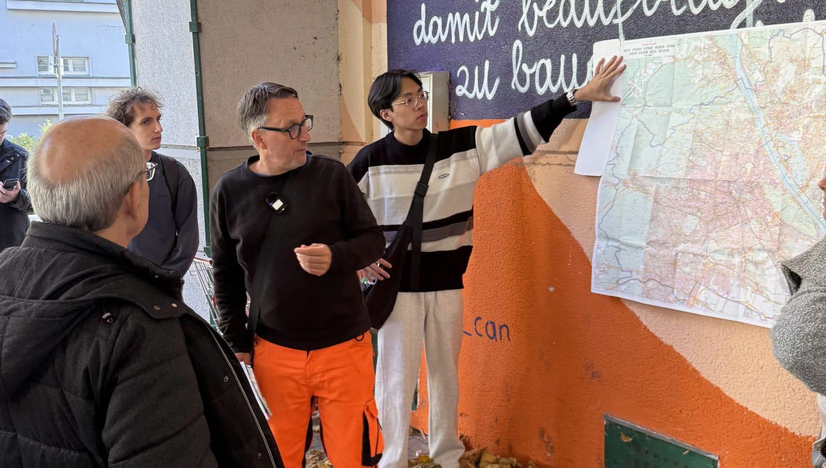 Eine Gruppe von Menschen steht draußen an einer Gebäudewand, die mit orangefarbener Farbe und Schrift bedeckt ist. Eine Person zeigt auf eine große Karte, die an die Wand geklebt ist, während andere zusehen und zuhören. Gefallene Blätter bedecken den Boden.