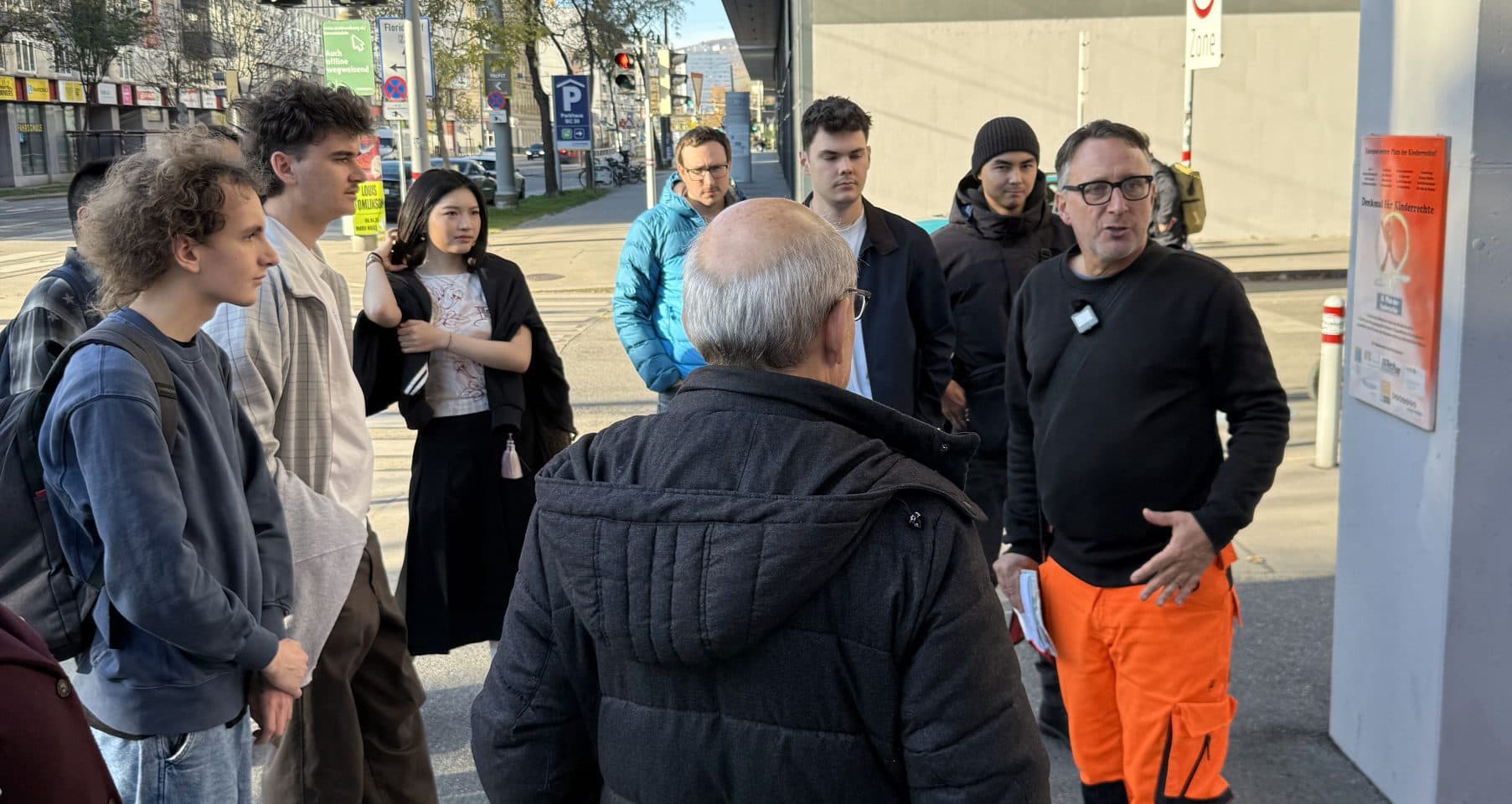 Eine Gruppe von Menschen versammelt sich um einen Mann in einer orangefarbenen Arbeitshose und einem schwarzen Hemd, der zu sprechen scheint oder Anweisungen gibt. Sie stehen auf einem Bürgersteig in der Nähe eines großen Betonbaus.