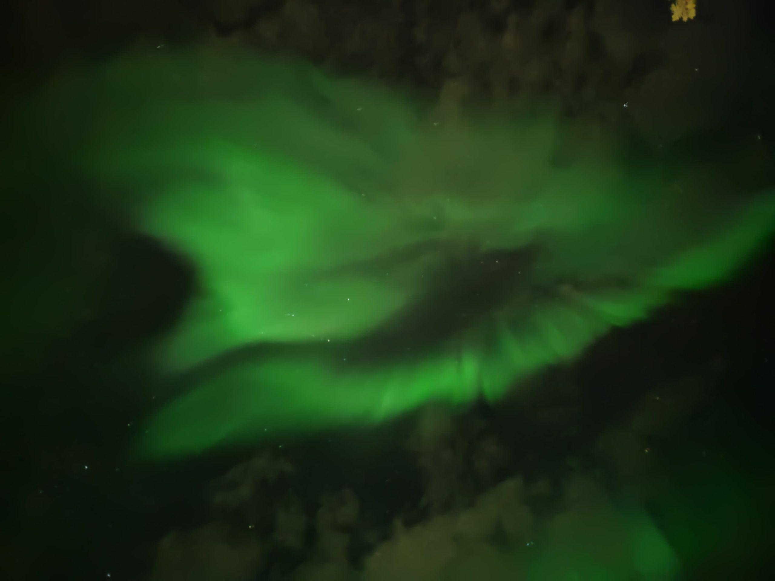 Grüne Polarlichter wirbeln über den dunklen Nachthimmel, und schwache Sterne sind hinter Wolkenfetzen sichtbar.