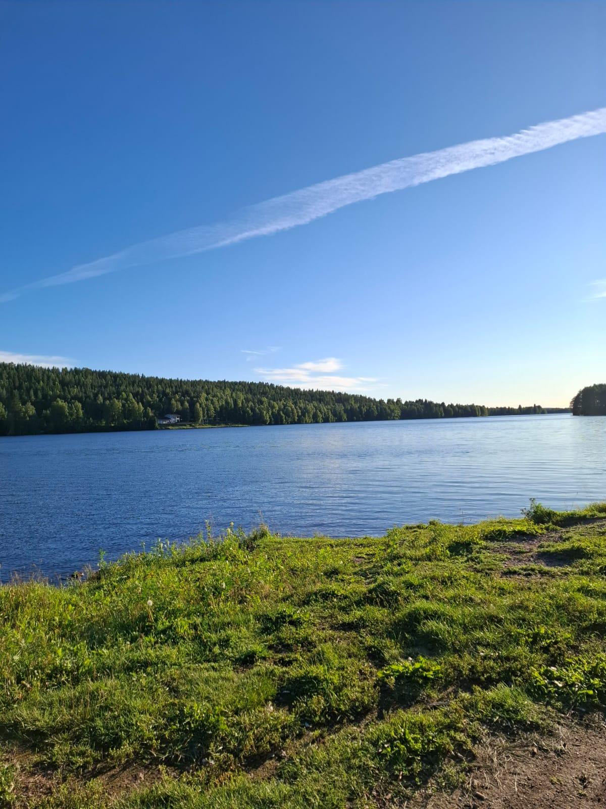 Ein ruhiger Fluss, gesäumt von einem grünen, grasbewachsenen Ufer und Bäumen, unter einem klaren, blauen Himmel, mit einer einzigen langen, weißen Wolke, die sich diagonal über den Himmel zieht.