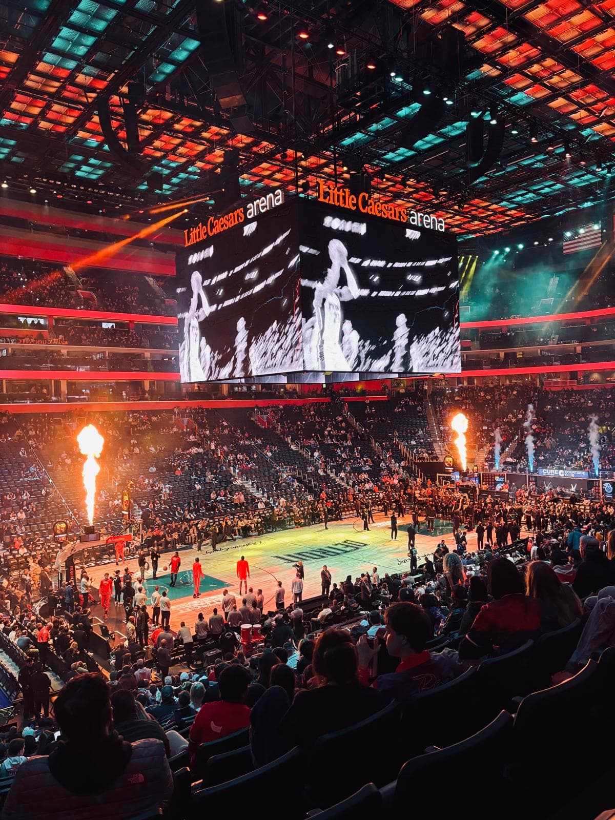 Eine vollbesetzte Basketball-Arena mit einer großen Anzeigetafel zeigt einen Spieler an; Flammen schießen auf dem Spielfeld während der Einleitungen vor dem Spiel in der Little Caesars Arena in die Höhe, während die Fans von den Tribünen aus zusehen.