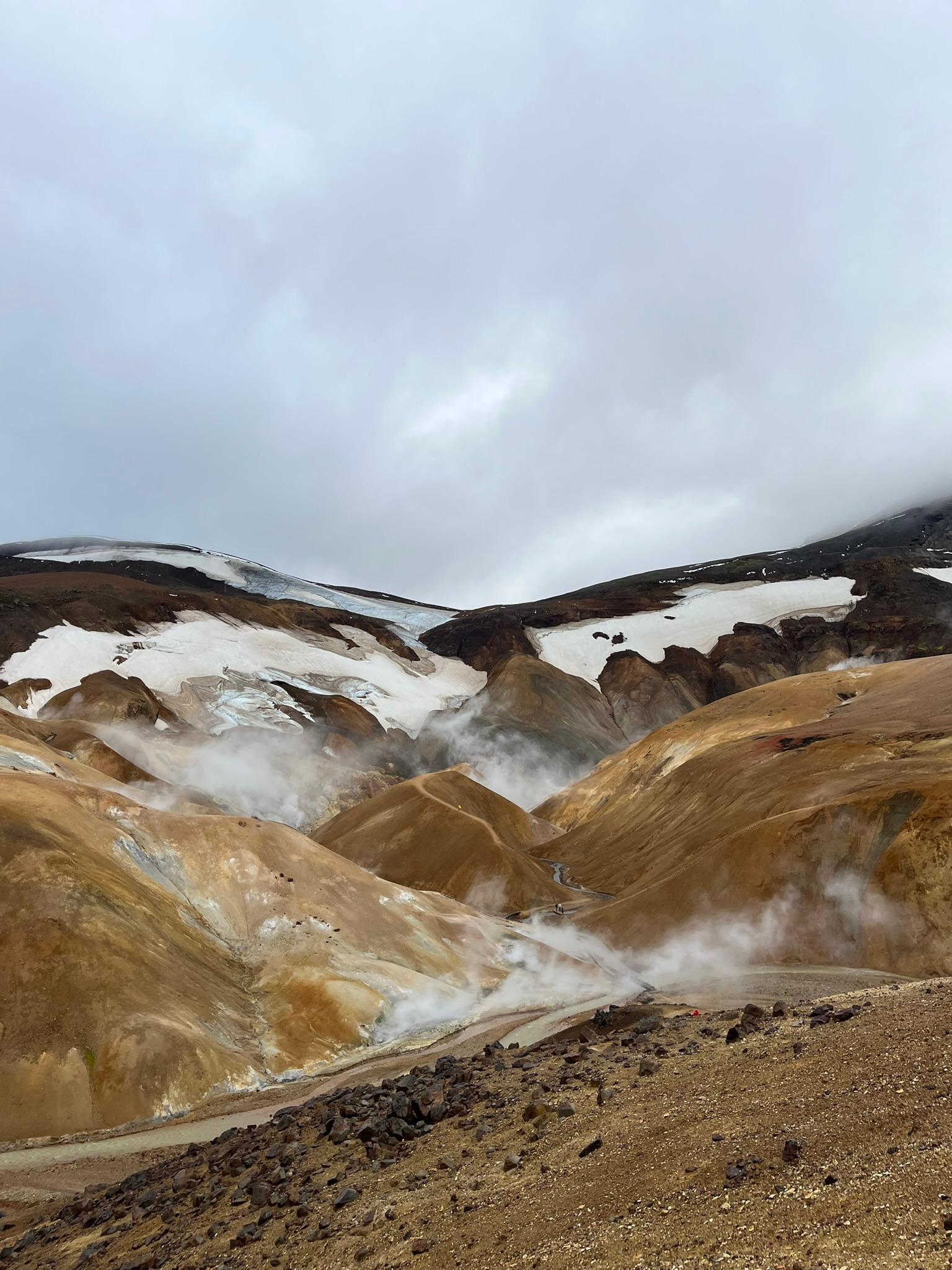Dampfende geothermische Schlote erheben sich aus ockerfarbenen und braunen Hügeln, die unter einem wolkenverhangenen Himmel mit Schneeflecken übersät sind und eine dramatische, raue Landschaft bilden.