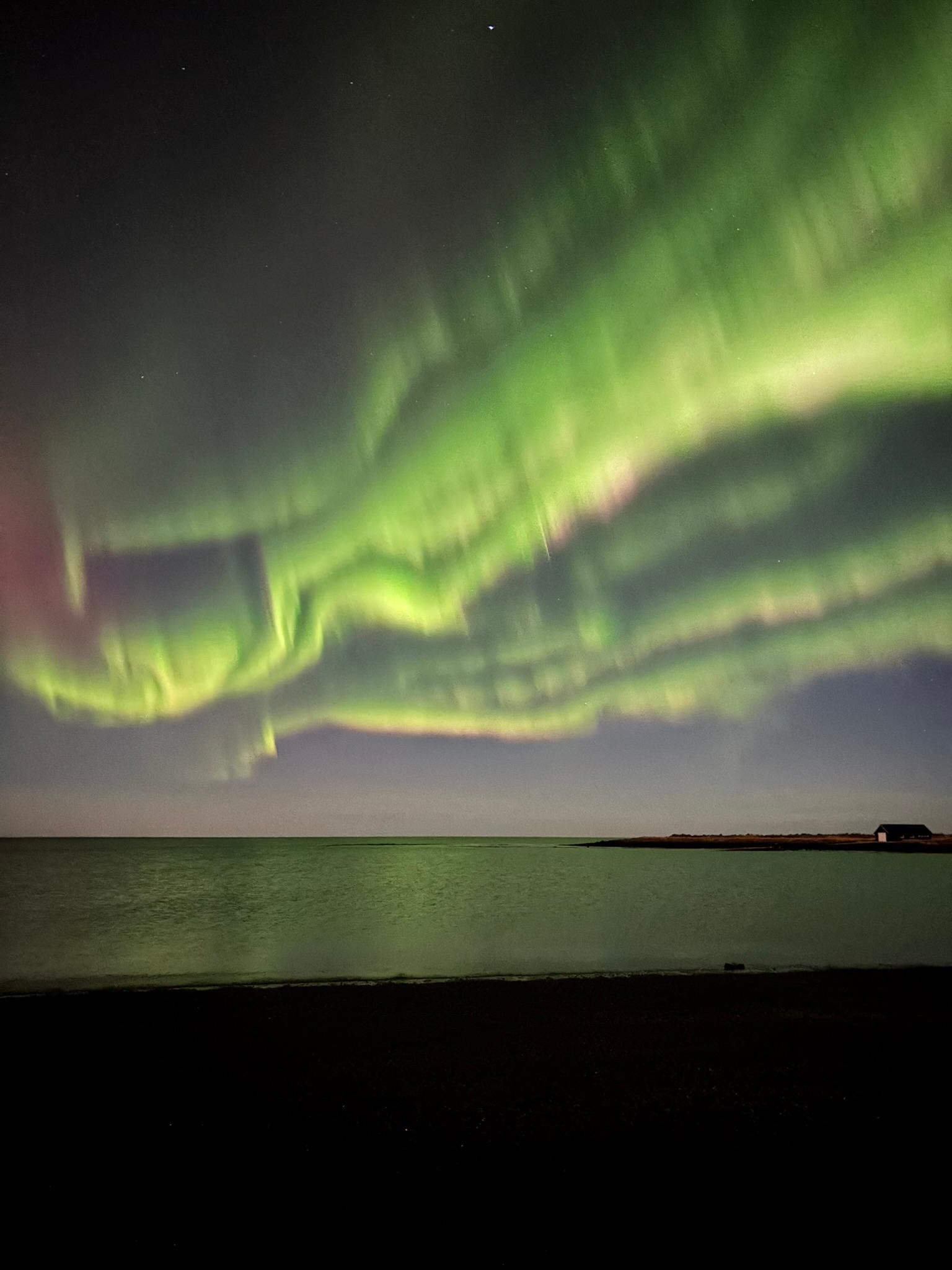 Grüne Polarlichter wirbeln über einen dunklen Nachthimmel über einem ruhigen Gewässer und einer entfernten Küstenlinie, mit einer schwachen Reflexion der Lichter auf der Wasseroberfläche.