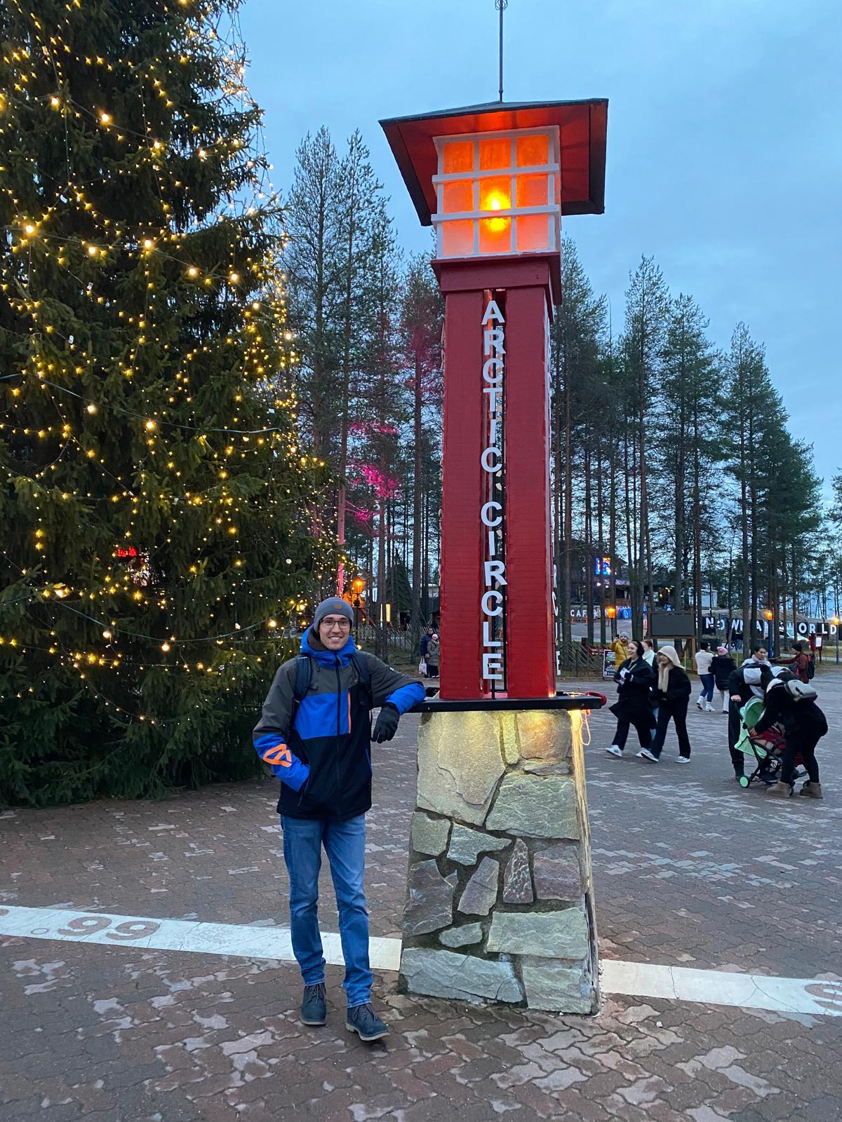 Eine Person in einer blauen Jacke steht neben einer roten Polarkreismarkierung mit einer leuchtenden Laterne an der Spitze, umgeben von Bäumen und dekorierten Lichtern, auf einem Platz im Freien mit Menschen im Hintergrund.