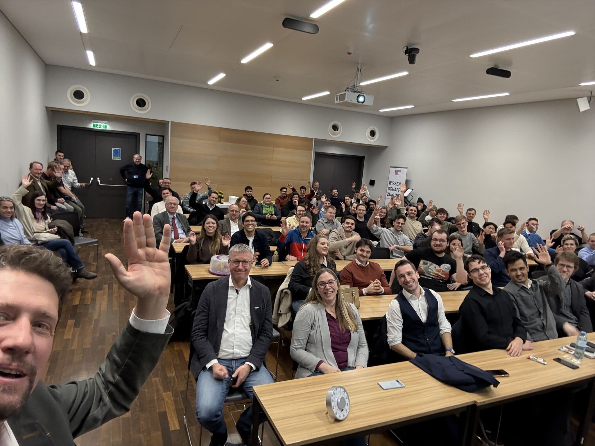 Eine große Gruppe von Menschen sitzt in Reihen mit Blick auf die Kamera in einem modernen Klassenzimmer oder Konferenzraum. Ein Mann im Vordergrund winkt, und die meisten Teilnehmer lächeln, was eine fröhliche, lebhafte Atmosphäre schafft.