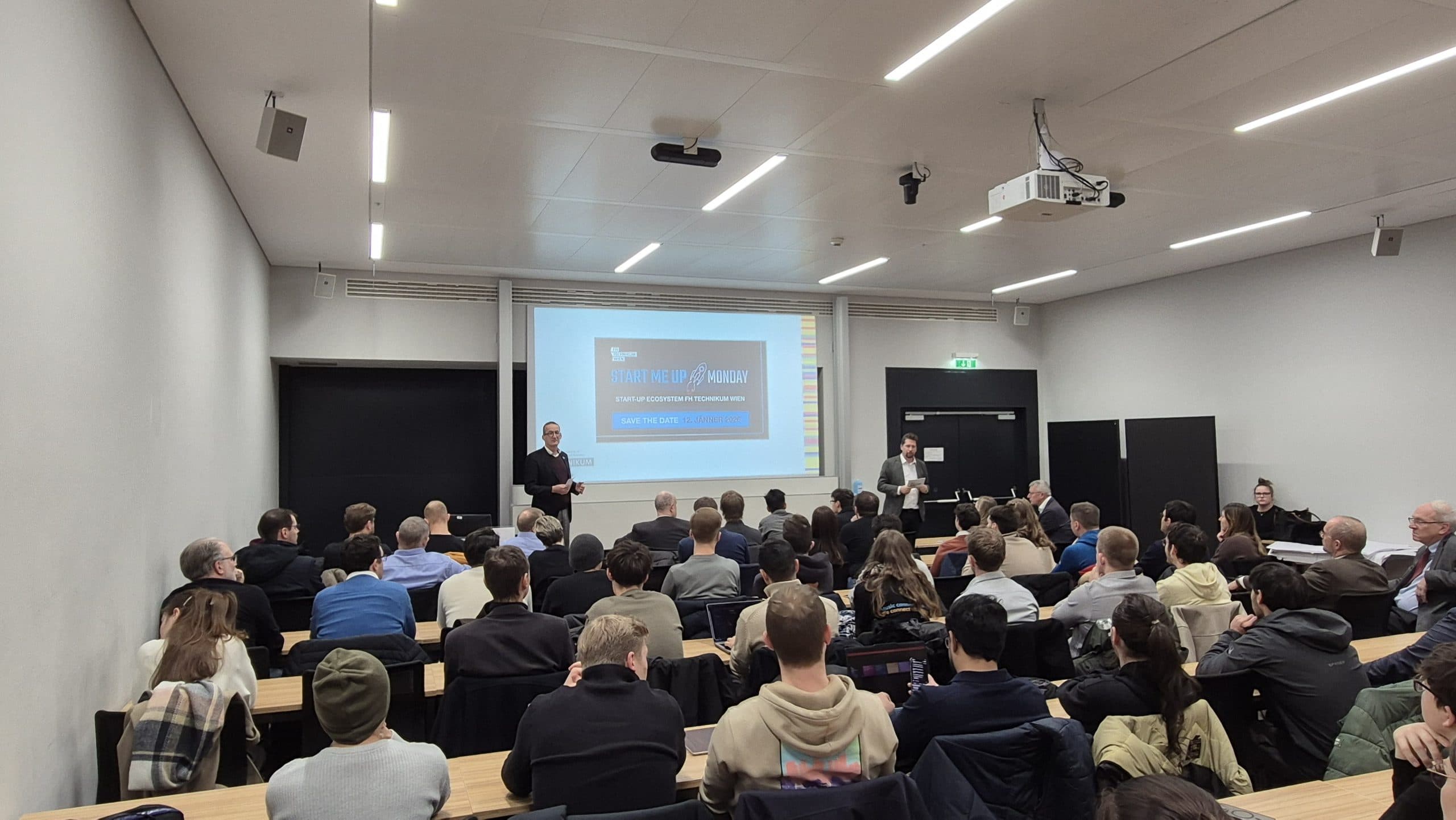 Ein Klassenzimmer mit sitzenden Teilnehmern, die auf einen Bildschirm an der Stirnseite blicken, wo zwei Referenten stehen und einen Vortrag mit dem Titel Startup M&As Unpacked halten. Der Raum ist hell erleuchtet und modern eingerichtet.