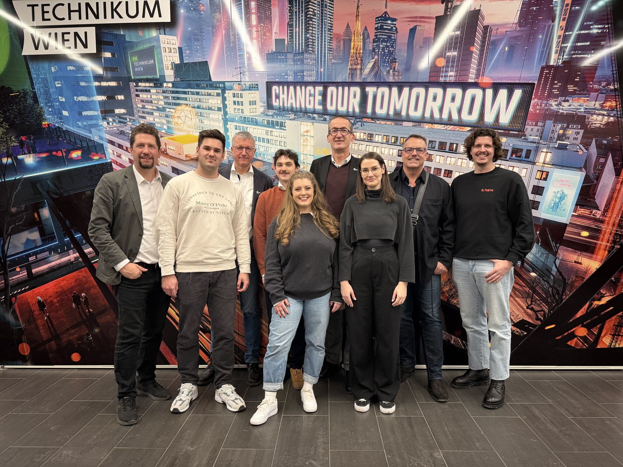 Eine Gruppe von neun Personen steht lächelnd vor einem farbenfrohen Wandbild mit städtischen Gebäuden und dem Text 'CHANGE OUR TOMORROW' im Technikum Wien.