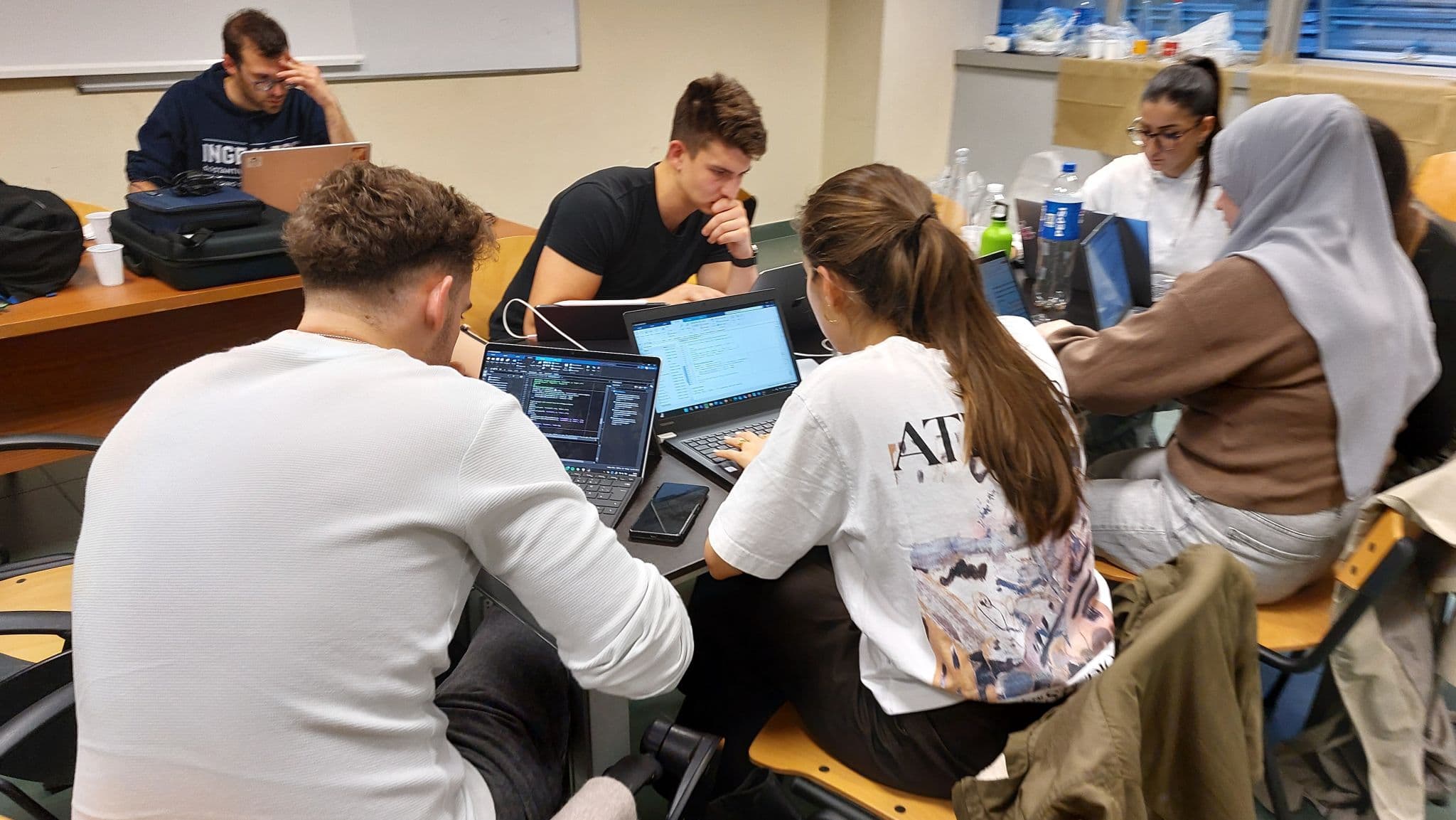 Fünf Schüler sitzen an Tischen in einem Klassenzimmer und arbeiten an Laptops. Drei von ihnen sind mit Programmier- oder technischen Aufgaben beschäftigt, und zwei arbeiten an einem anderen Tisch im Hintergrund. Auf den Tischen stehen Flaschen und Taschen.