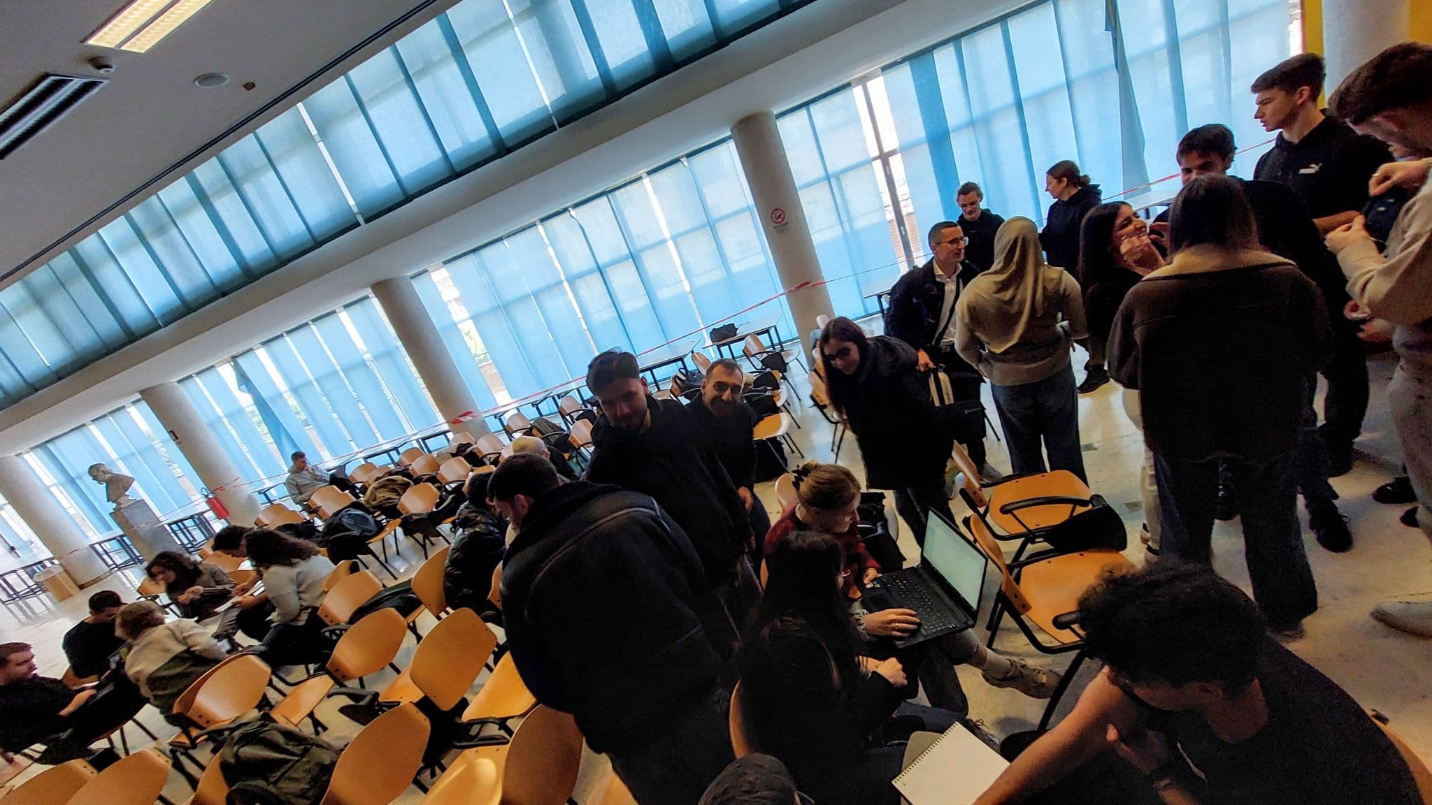In einem geräumigen Klassenzimmer mit vielen leeren Stühlen und großen blau getönten Fenstern versammelt sich eine Gruppe von Schülern und plaudert, während einige an ihren Laptops arbeiten.