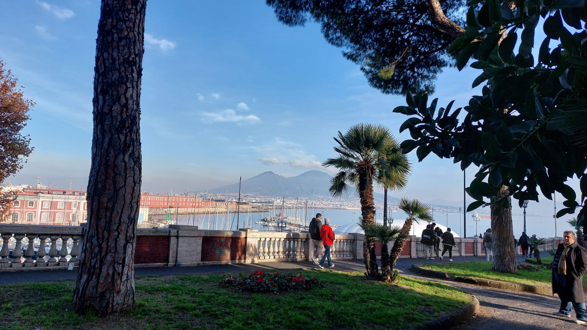 Spaziergänger in einem Park mit Bäumen und Blumenbeeten, mit Blick auf einen Hafen mit Booten. Im Hintergrund erhebt sich der Vesuv unter einem blauen Himmel mit vereinzelten Wolken.