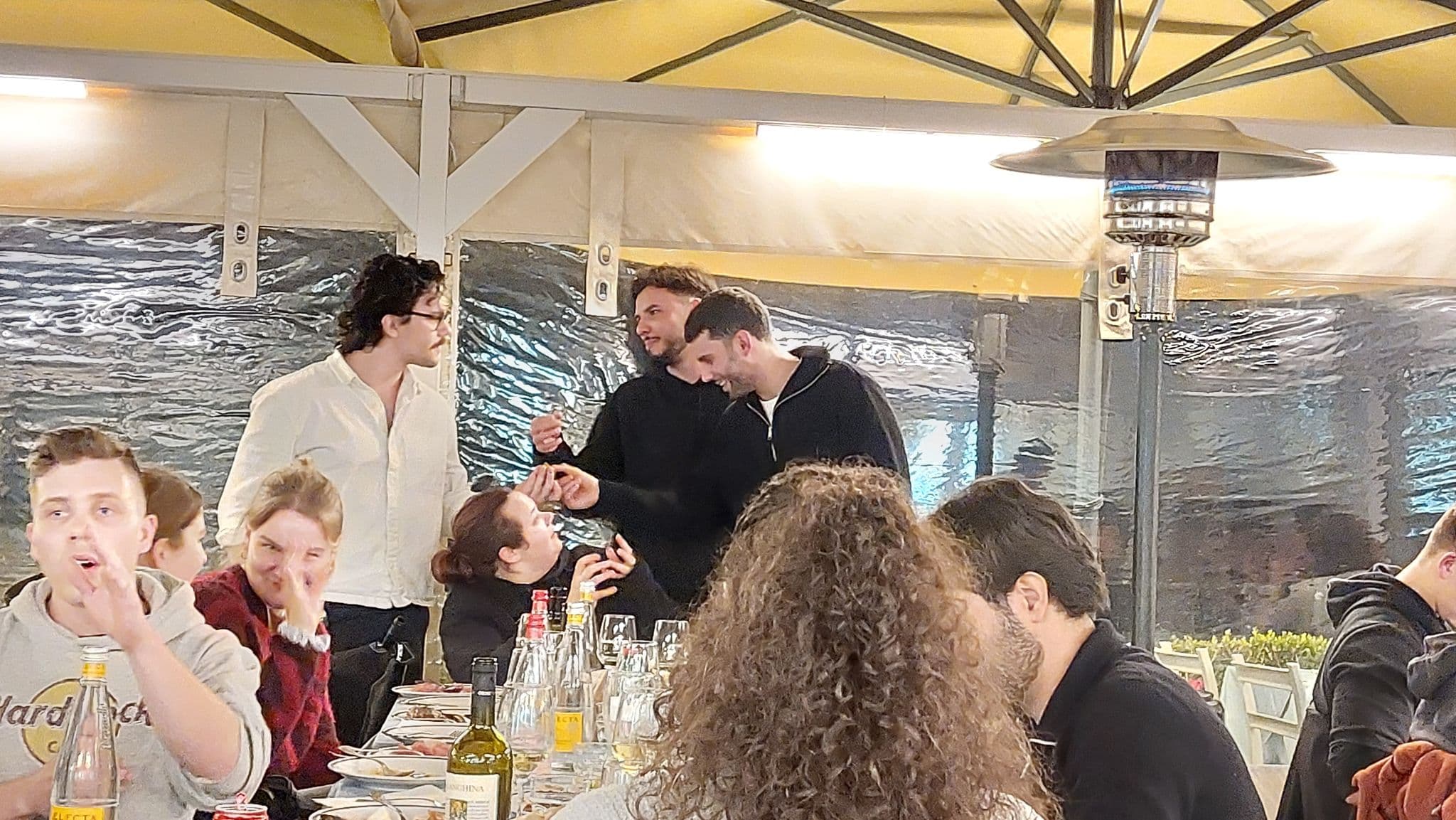 Eine Gruppe von Menschen ist um einen langen Tisch in einem Restaurant im Freien versammelt. Einige sitzen, essen und unterhalten sich, andere stehen und lachen zusammen. Die Atmosphäre ist lebhaft und gesellig.
