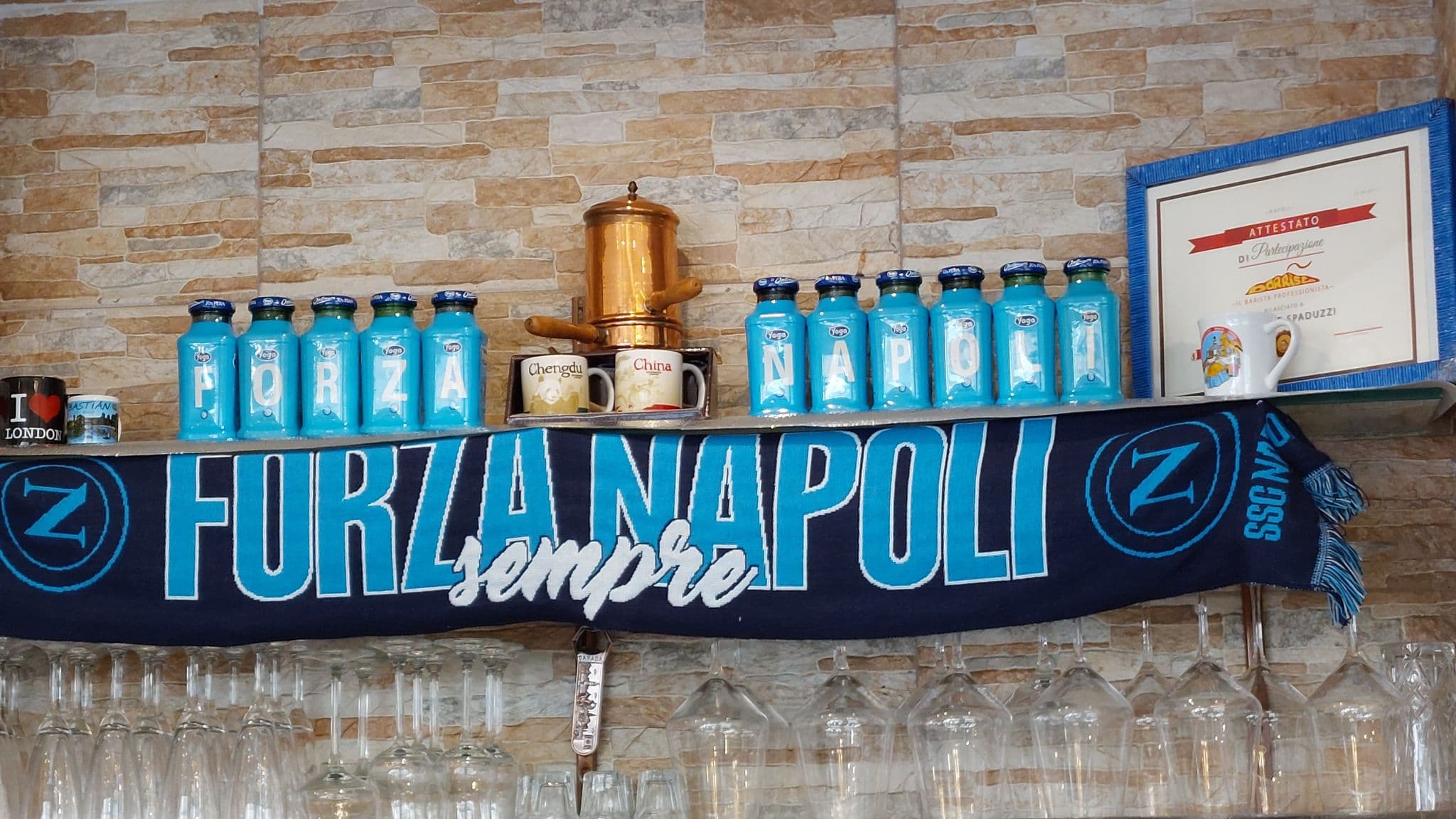 Auf einem Regal stehen blaue Gläser mit der Aufschrift 'FORZA NAPOLI', ein Napoli-Fußballschal, eine goldene Kaffeemaschine, kopfüber hängende Gläser und eine gerahmte Urkunde an einer Steinwand, die den Fußballverein Napoli feiert.