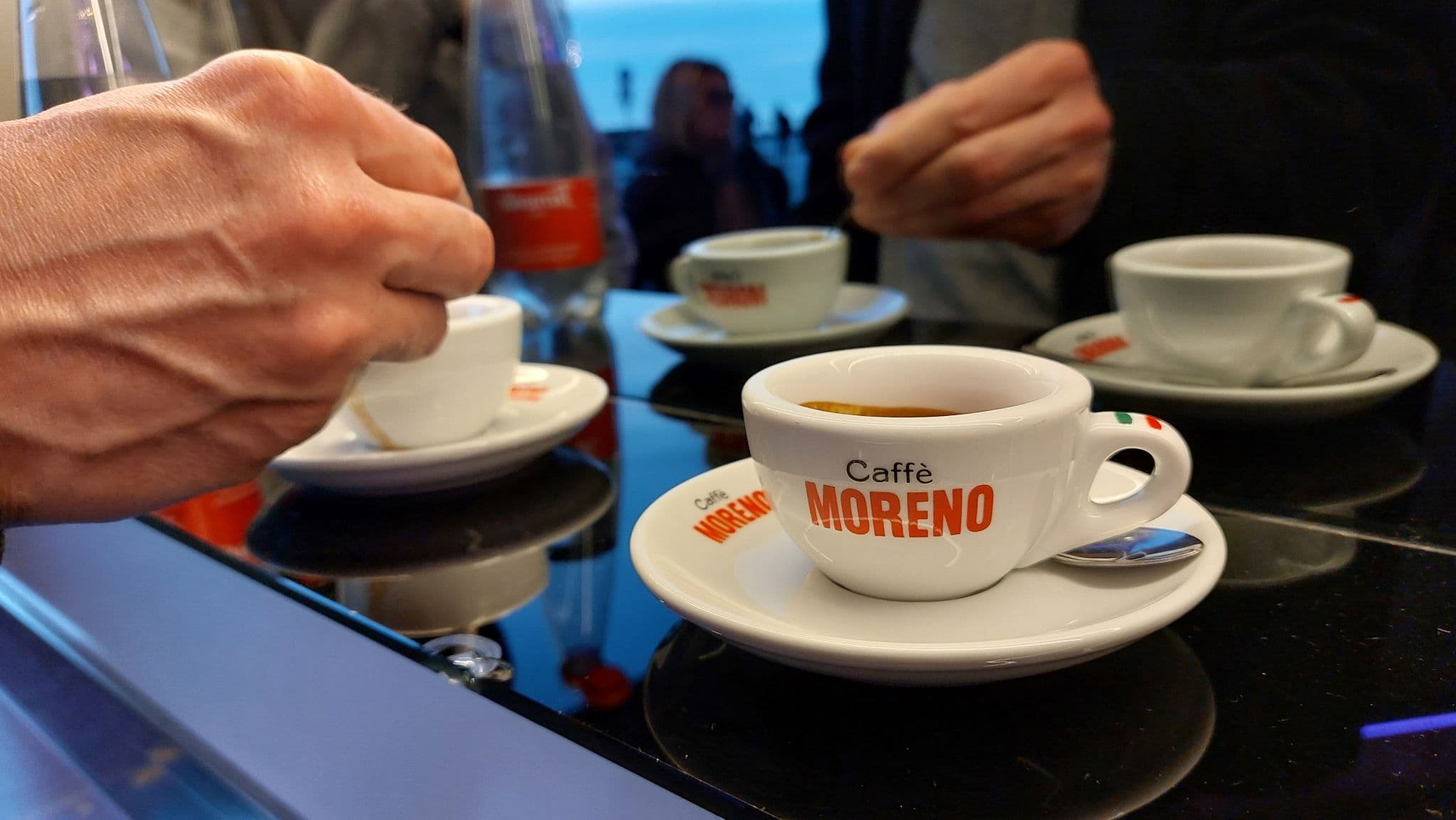 Nahaufnahme einer Hand, die eine Tasse Espresso auf einer Untertasse mit der Aufschrift Caffè Moreno umrührt, mit einer weiteren Tasse und einer Flasche im Hintergrund, die sich in einer glänzenden schwarzen Oberfläche spiegeln.