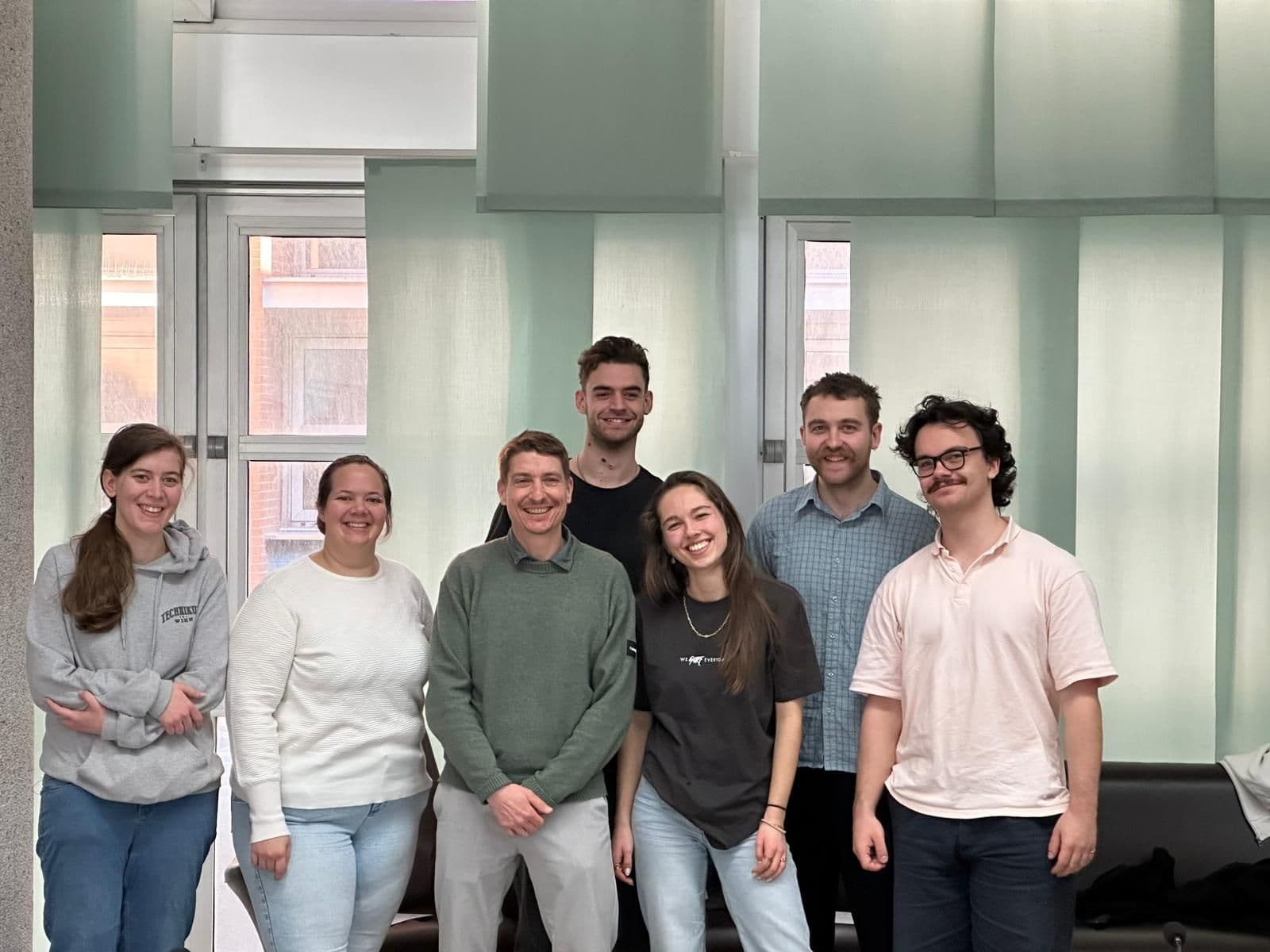 Sieben Personen stehen in einem Innenraum vor großen Fenstern mit grünen Jalousien und lächeln in die Kamera. Sie sind leger gekleidet und in einer lockeren Reihe angeordnet. Die Atmosphäre wirkt freundlich und entspannt.