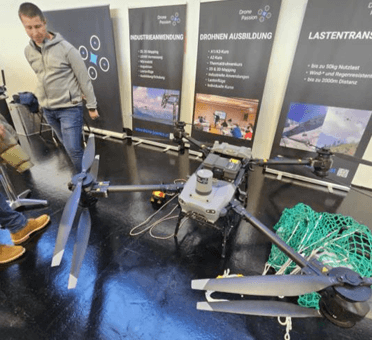 Ein Mann steht neben einer großen Industriedrohne mit vier Propellern, die bei einer Indoor-Veranstaltung ausgestellt ist. Dahinter sind Banner zu sehen, die für Drohnenschulungen, industrielle Anwendungen und Frachttransport werben.