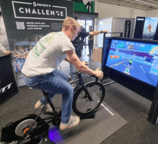 Ein Mann fährt auf einem stationären Fahrrad, das mit einem Bildschirm verbunden ist, auf dem ein virtuelles Radrennen an einem Indoor-Event-Stand angezeigt wird. Eine andere Person steht in der Nähe und interagiert mit dem Standaufbau.