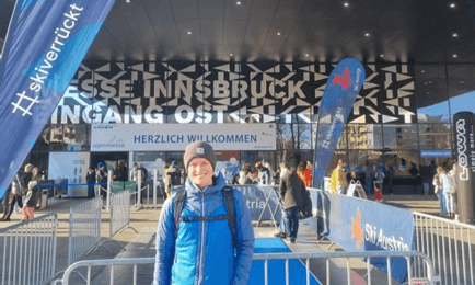 Eine lächelnde Person in blauer Jacke und Mütze steht vor der Messe Innsbruck, unter einem Vordach mit Herzlich Willkommen und Veranstaltungsbannern. Das Sonnenlicht wirft Schatten auf den Boden.