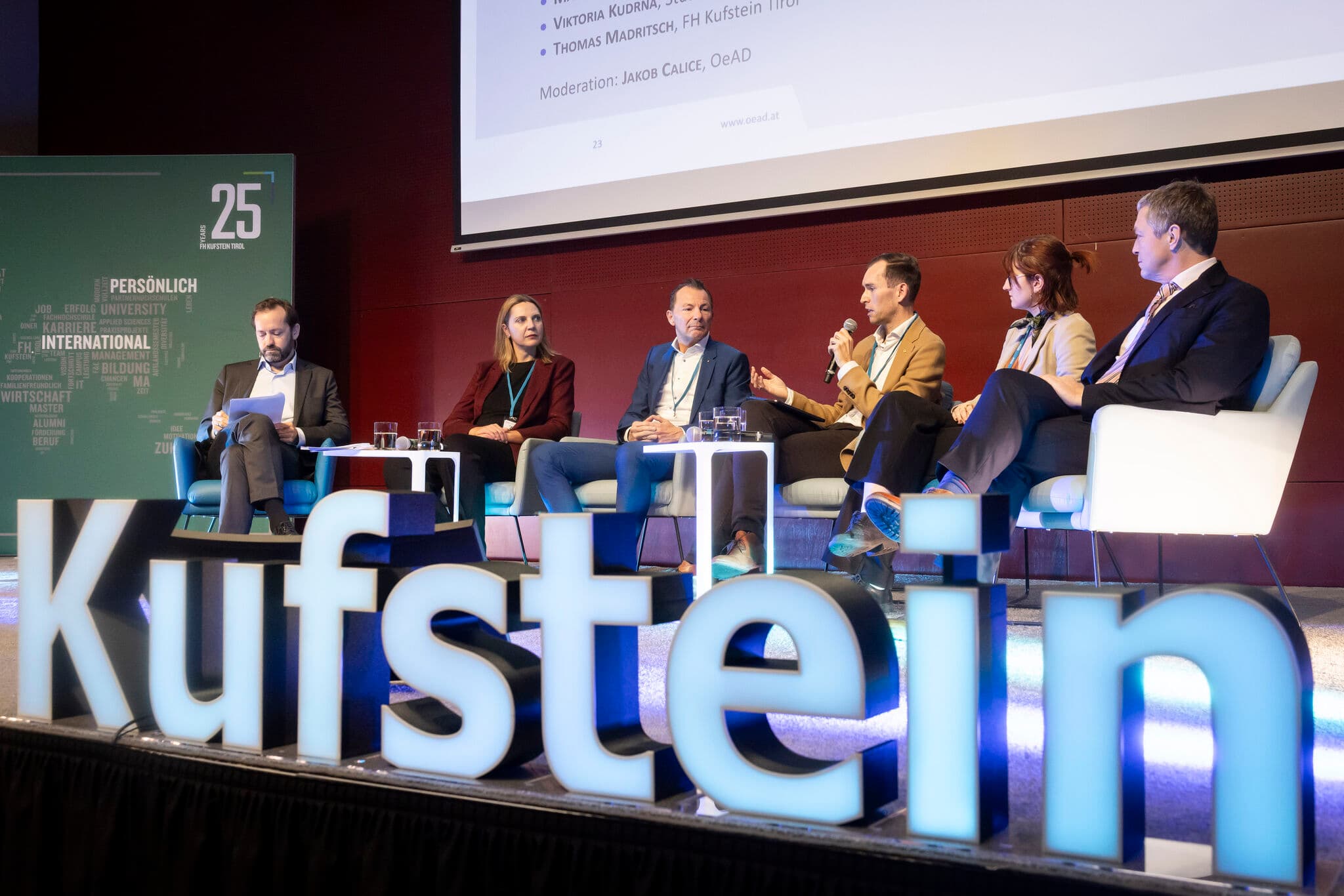 Sechs Personen sitzen auf einer Bühne in einer Podiumsdiskussion, mit Mikrofonen und einem sichtbaren Moderator. Im Vordergrund steht ein großes beleuchtetes Schild mit der Aufschrift Kufstein, und dahinter ist eine Präsentation zu sehen.