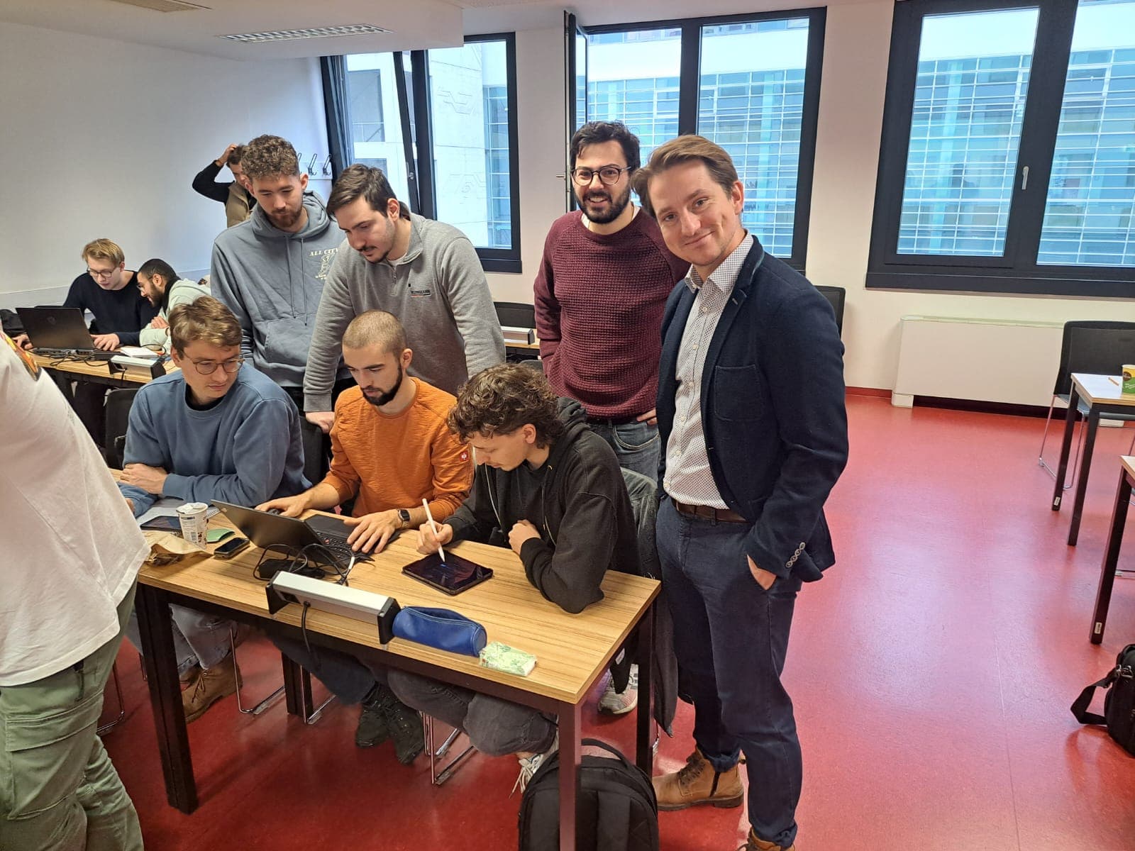 Eine Gruppe junger Erwachsener, die um einen Schreibtisch in einem Klassenzimmer versammelt sind. Einige sitzen und arbeiten an Laptops, während andere lächelnd auf den Bildschirm schauen. Im Hintergrund sind große Fenster und weitere Schüler zu sehen.