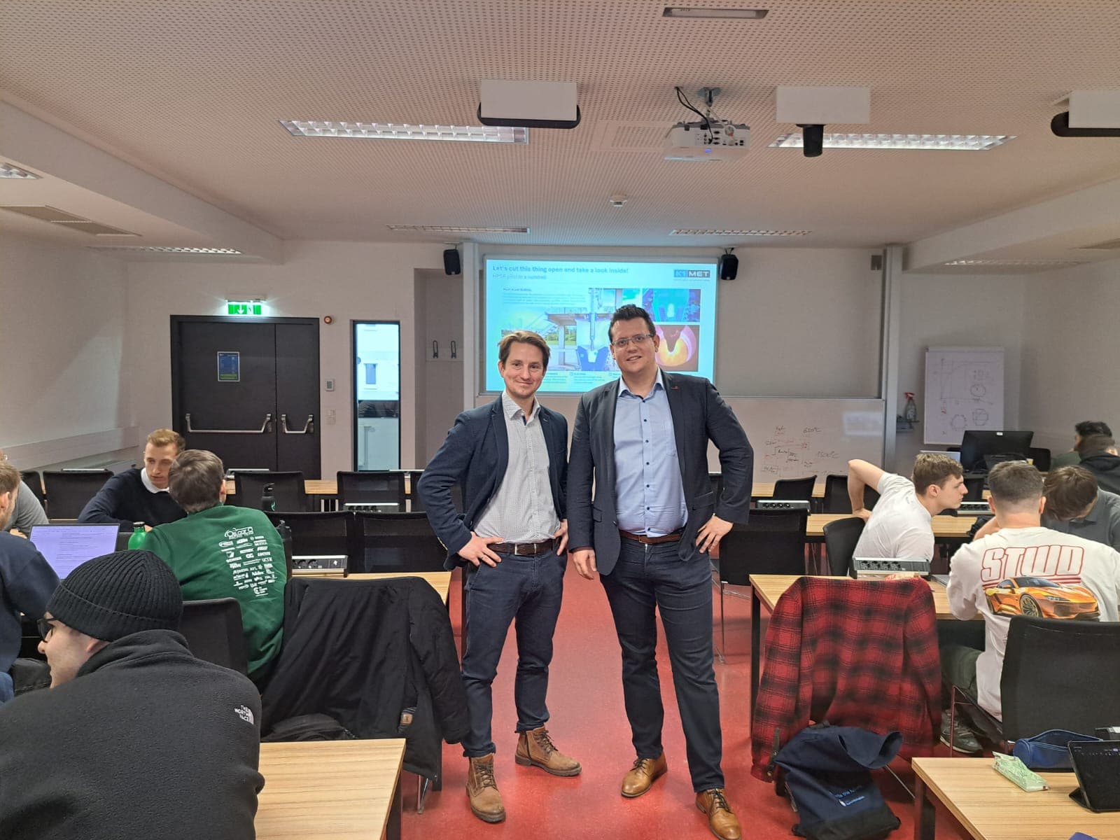 Zwei Männer in Geschäftskleidung stehen lächelnd vor einem Klassenzimmer, in dem Schüler mit Laptops an den Tischen sitzen. Hinter ihnen wird eine Präsentation auf die Leinwand projiziert. Die Umgebung ist hell und modern.
