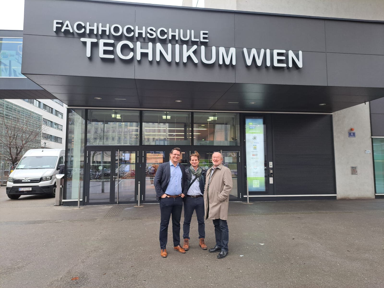 Drei Männer stehen lächelnd vor dem Haupteingang der Fachhochschule Technikum Wien, einer technischen Universität in Wien. Das Gebäude hat große Glastüren und ein markantes Schild über dem Eingang.