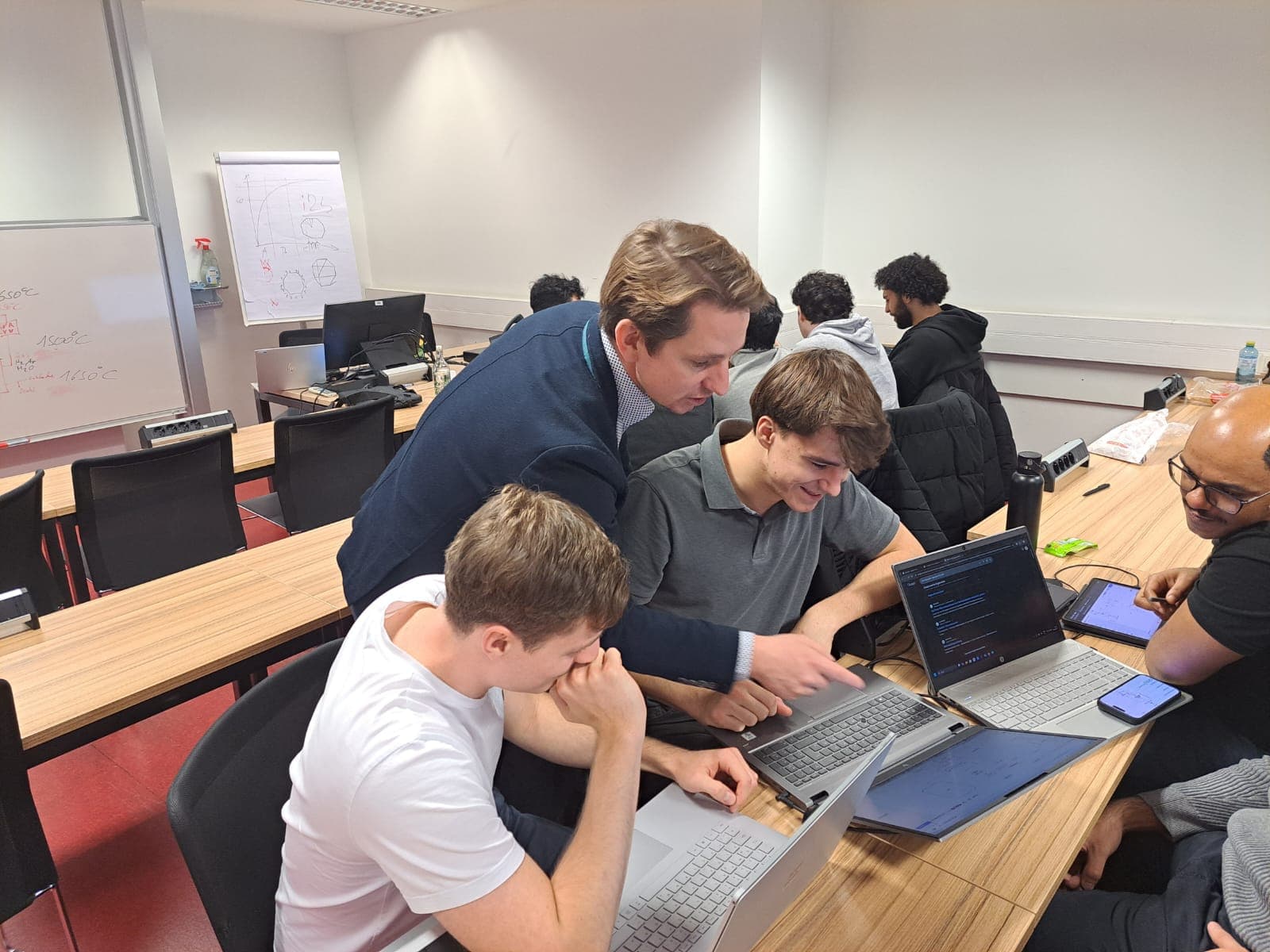 Ein Lehrer hilft zwei Schülern in einem Klassenzimmer bei der Programmierung auf Laptops, wobei er auf einen Bildschirm zeigt, während eine andere Gruppe im Hintergrund arbeitet. Ein Whiteboard mit Diagrammen ist zu sehen.