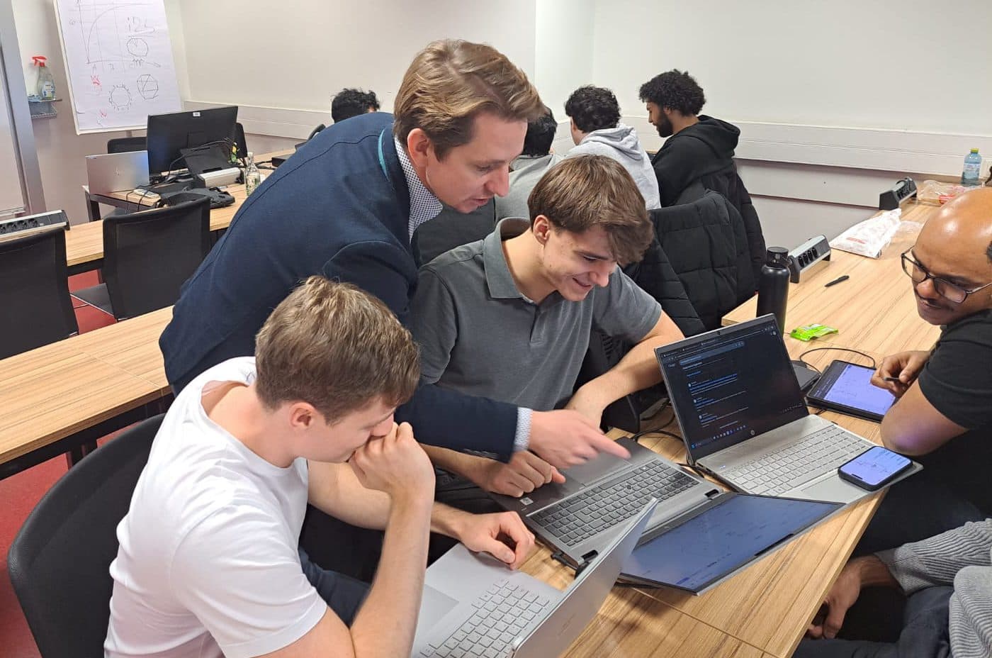 Ein Mann im Anzug beugt sich über zwei sitzende Studenten und zeigt auf einen Laptop-Bildschirm. Die Schüler sehen engagiert aus und arbeiten an ihren Laptops in einem Klassenzimmer mit anderen Schülern und einer Tafel im Hintergrund.