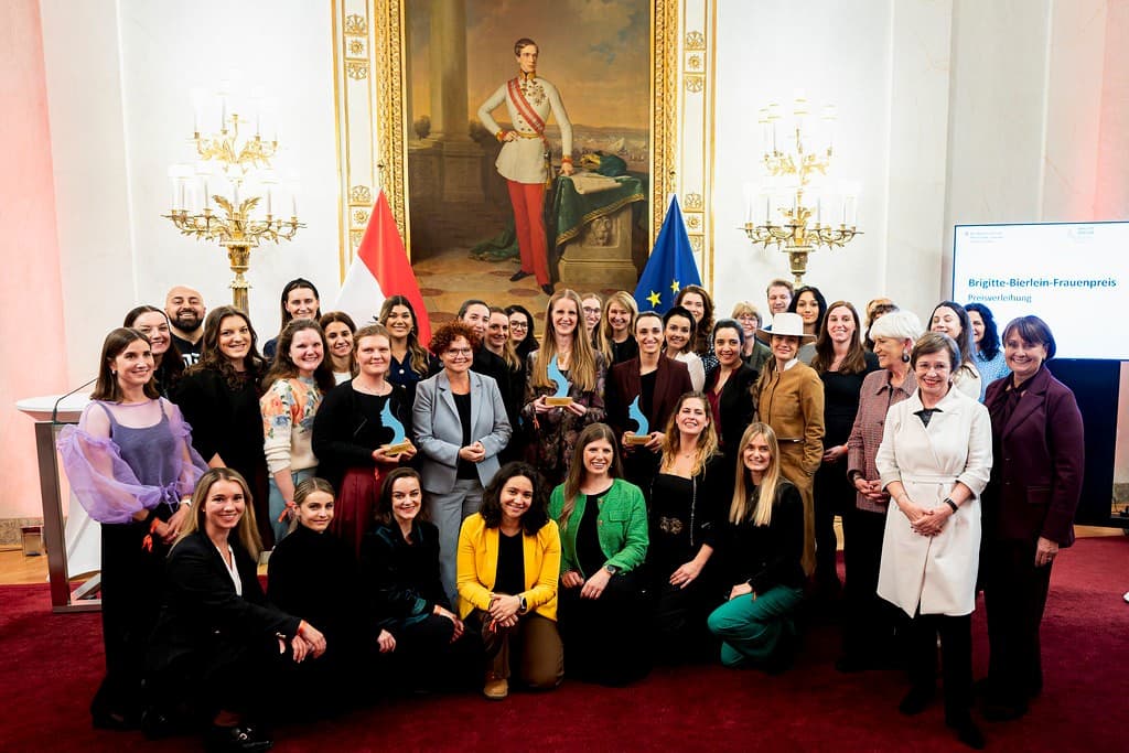 Eine große Gruppe von Menschen, zumeist Frauen, posiert lächelnd in einem prächtig dekorierten Raum mit einem großen Gemälde eines Mannes in Uniform, Fahnen und Kronleuchtern. Einige halten Trophäen oder Auszeichnungen, was auf eine feierliche Veranstaltung hindeutet.