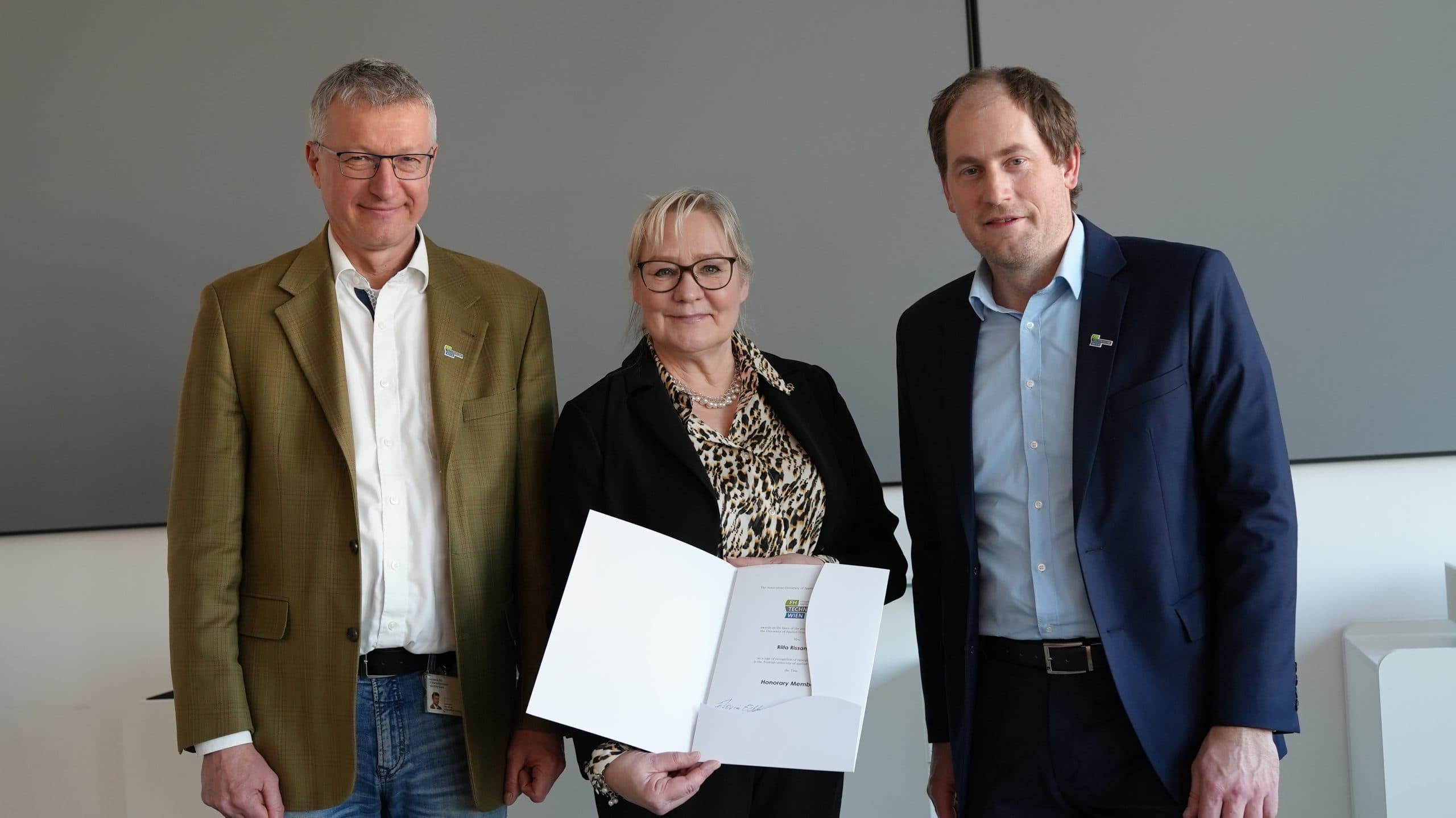 Drei Personen stehen in einem Haus und lächeln in die Kamera. Die Frau in der Mitte hält eine offene Urkunde in der Hand, flankiert von zwei Männern in Business-Kleidung. Der Hintergrund besteht aus schlichten, grauen Tafeln.