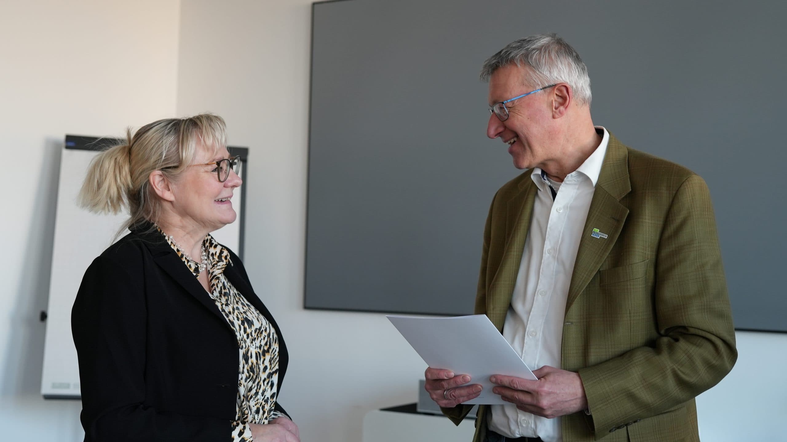 Eine Frau und ein Mann stehen in einem Haus und lächeln sich an. Der Mann hält ein Dokument in der Hand. Beide sind in Geschäftskleidung gekleidet, und im Hintergrund sind ein Flipchart und eine große graue Tafel zu sehen.