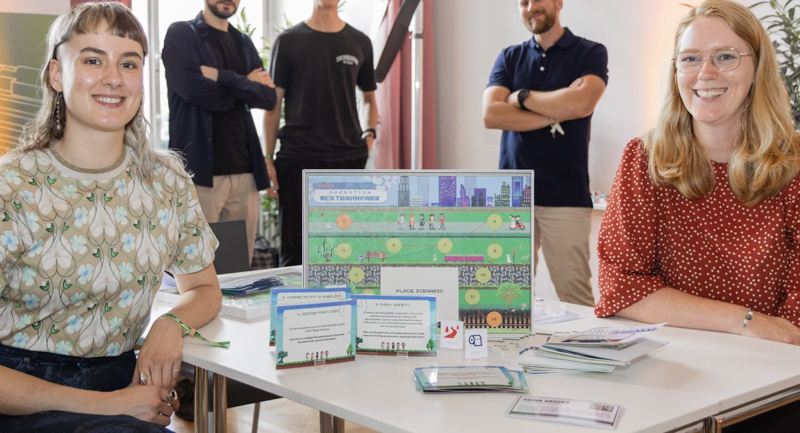 Zwei Frauen sitzen an einem Tisch, der ein buntes Brettspiel mit Karten und Broschüren zeigt. Drei Personen stehen lächelnd im Hintergrund. Die Szene scheint auf einer Veranstaltung oder Messe zu spielen, mit einer hellen und einladenden Atmosphäre.