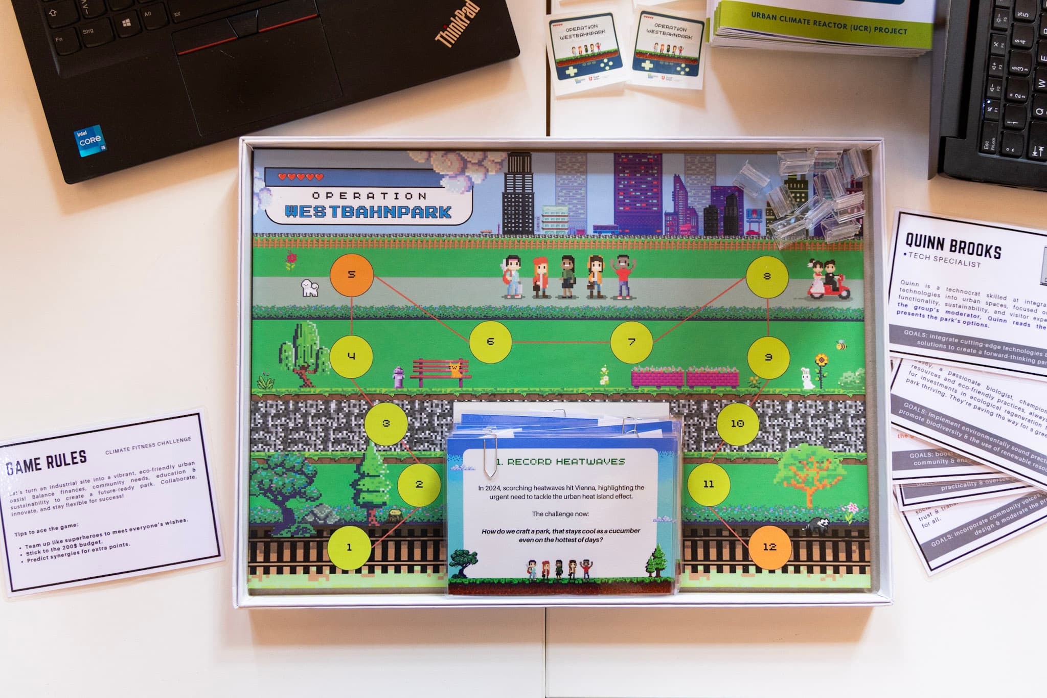 Ein farbenfrohes Brettspiel mit dem Namen 'Operation Westbahnpark' ist auf einem Tisch aufgebaut, mit einem nummerierten Pfad, Figurenplättchen, Karten und gedruckten Spielregeln. Laptops und Papiere sind um das Spiel herum zu sehen.