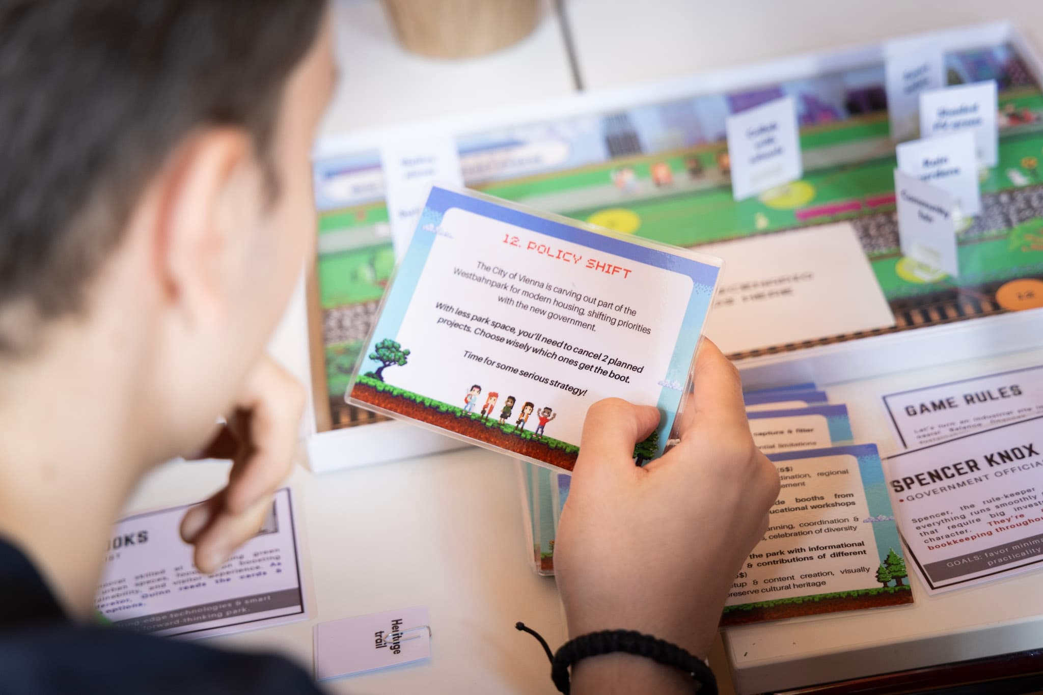 Eine Person hält eine Karte mit der Aufschrift 12. POLICY SHIFT und spielt ein Brettspiel. Das Spielbrett und verschiedene Karten mit Text und Illustrationen sind auf dem Tisch vor ihnen zu sehen.