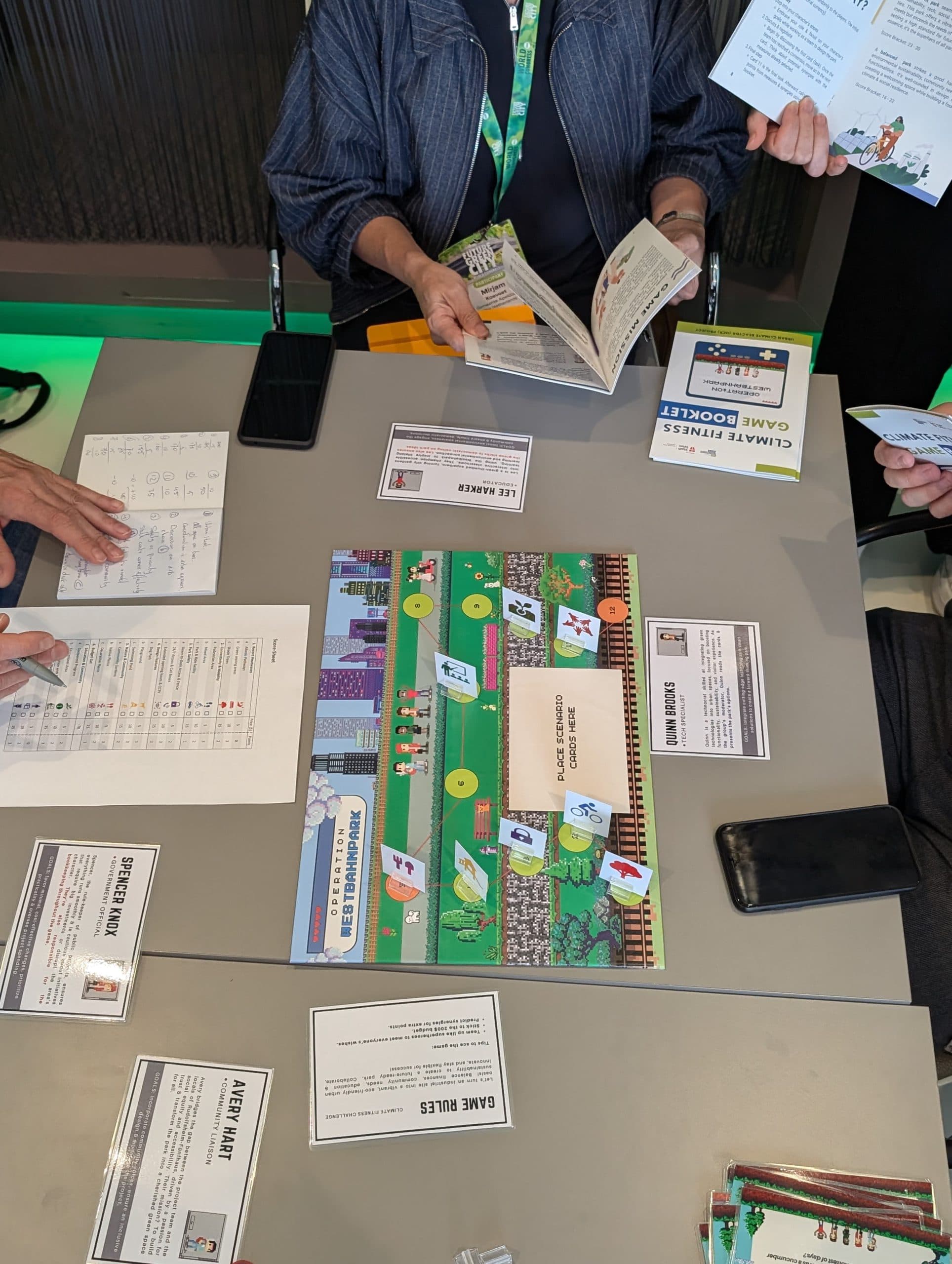 Drei Personen sitzen um einen Tisch und spielen ein Brettspiel namens Climate Fresk Game Board. Der Tisch ist mit Karten, Papieren und Mobiltelefonen bedeckt, während das Spielbrett bunte Grafiken und Spielanweisungen zeigt.