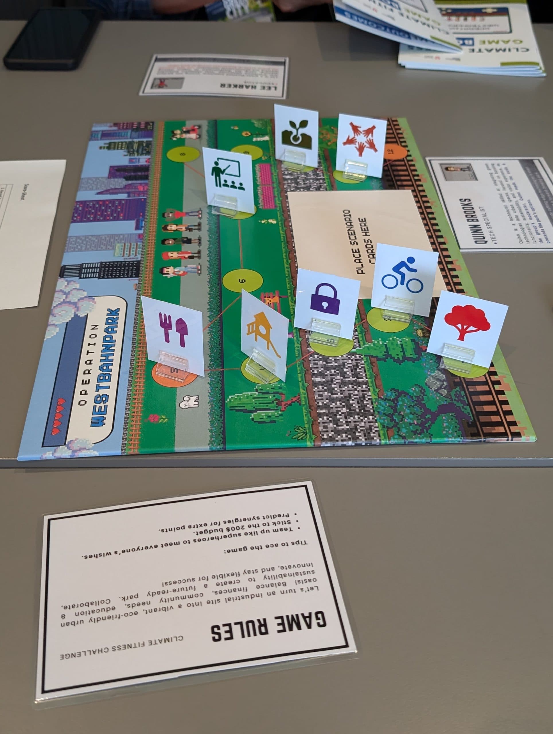 Auf einem Tisch liegt ein Brettspiel mit dem Titel 'Operation West Branch Park', auf dem Karten mit Symbolen für Bäume, Fahrräder, Lebensmittel, Wasser und Recycling abgebildet sind. In der Nähe befinden sich gedruckte Spielregeln und Papiermaterialien.
