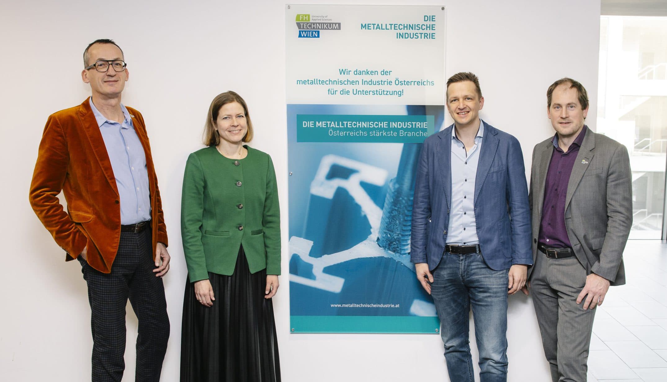 Vier Personen stehen in einem Raum vor einer weißen Wand mit einem blau-grünen Informationsplakat über die Metallindustrie. Sie lächeln und tragen Business-Casual-Kleidung.
