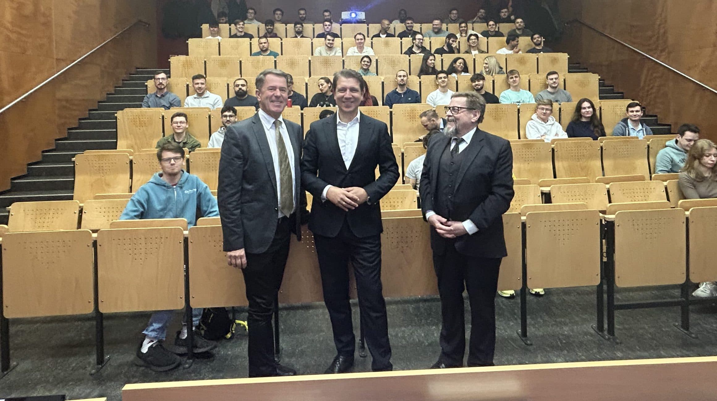 Drei Männer in Anzügen stehen am Anfang eines Hörsaals und lächeln in die Kamera. Hinter ihnen sitzt eine Gruppe von Studenten auf hölzernen Stühlen, die nach vorne gerichtet sind. Der Raum hat braune Wände und eine helle Deckenbeleuchtung.