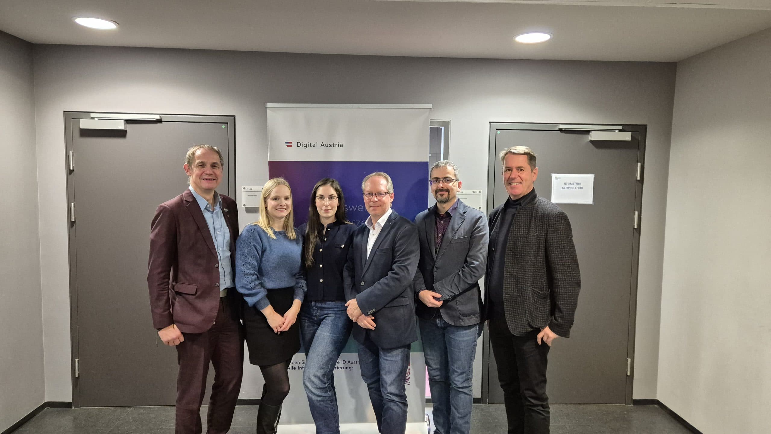 Sechs Personen stehen lächelnd nebeneinander vor einem Banner von Digital Austria mit zwei grauen Türen im Hintergrund. Die Gruppe besteht aus drei Männern in Anzügen, einem Mann in Jeans und zwei Frauen in legerer Business-Kleidung.