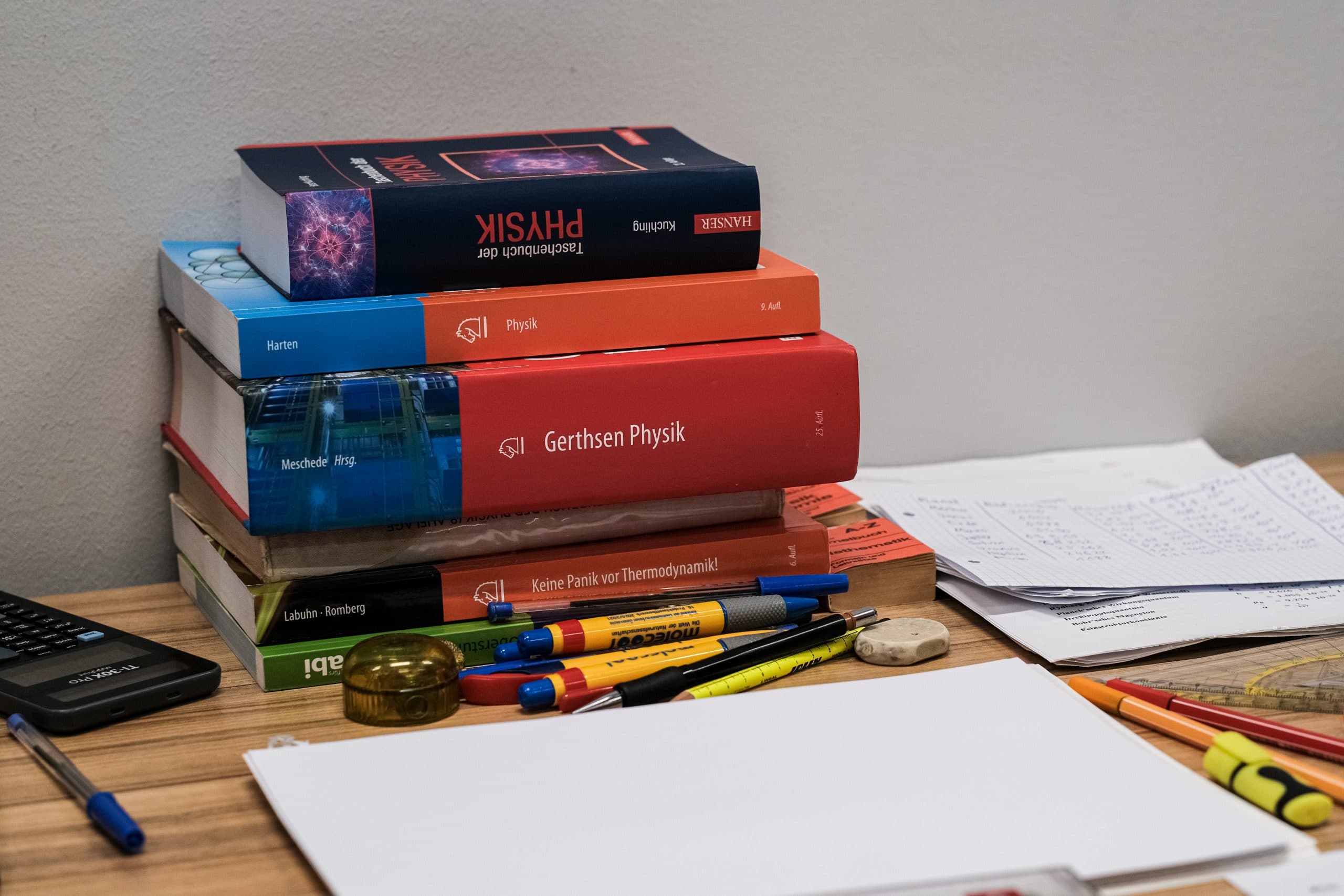 Auf einem hölzernen Schreibtisch liegen ein Stapel Physiklehrbücher und -hefte neben Textmarkern, Stiften, einem Taschenrechner, losen Papieren und einem Winkelmesser, was auf eine Lern- oder Hausaufgabenstunde schließen lässt.