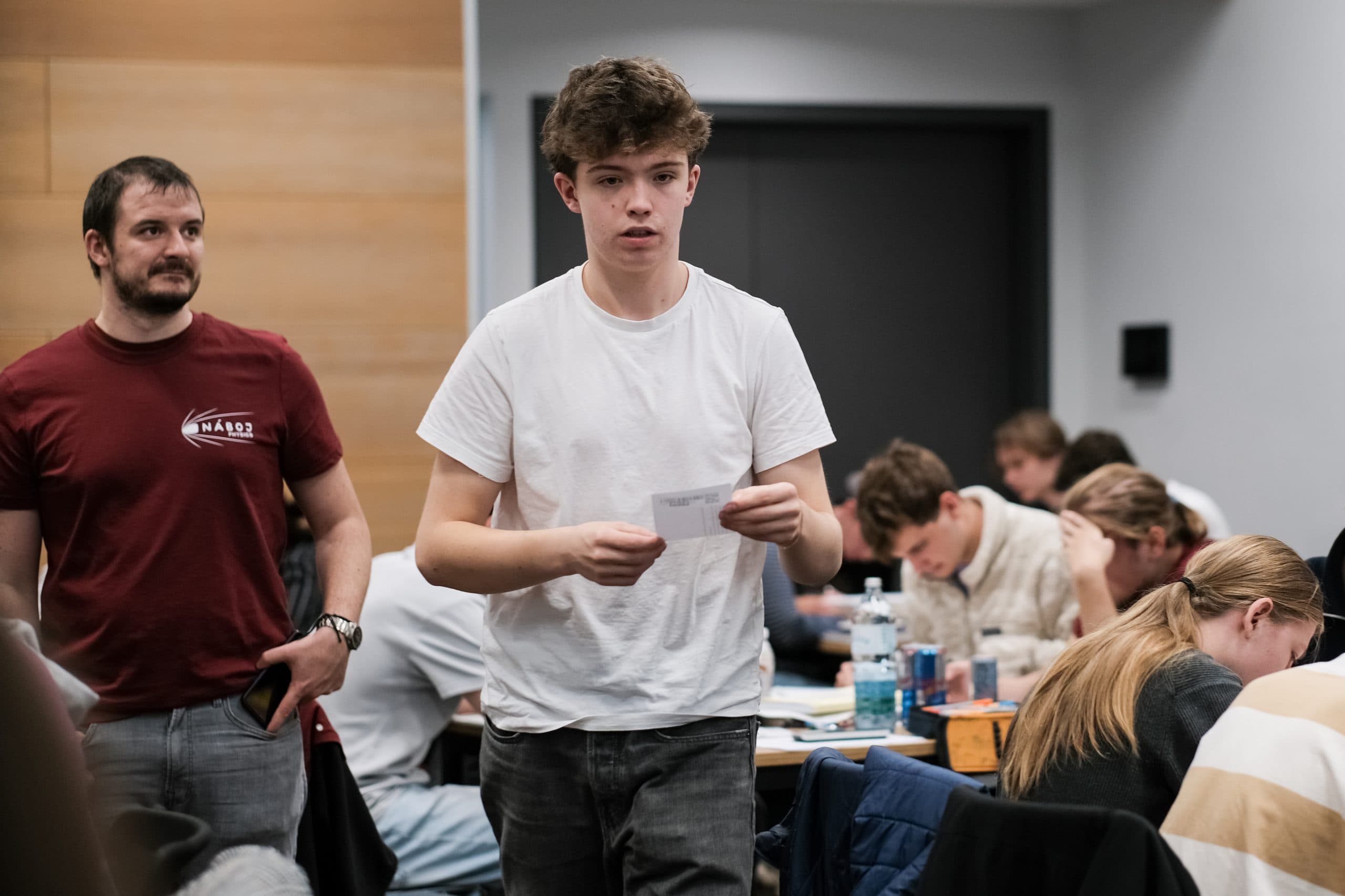 Ein junger Mann in einem weißen T-Shirt geht in einem belebten Klassenzimmer spazieren und hält eine Zeitung in der Hand. Im Hintergrund sind andere Schüler zu sehen, von denen einige an Tischen sitzen und arbeiten und ein Mann steht.