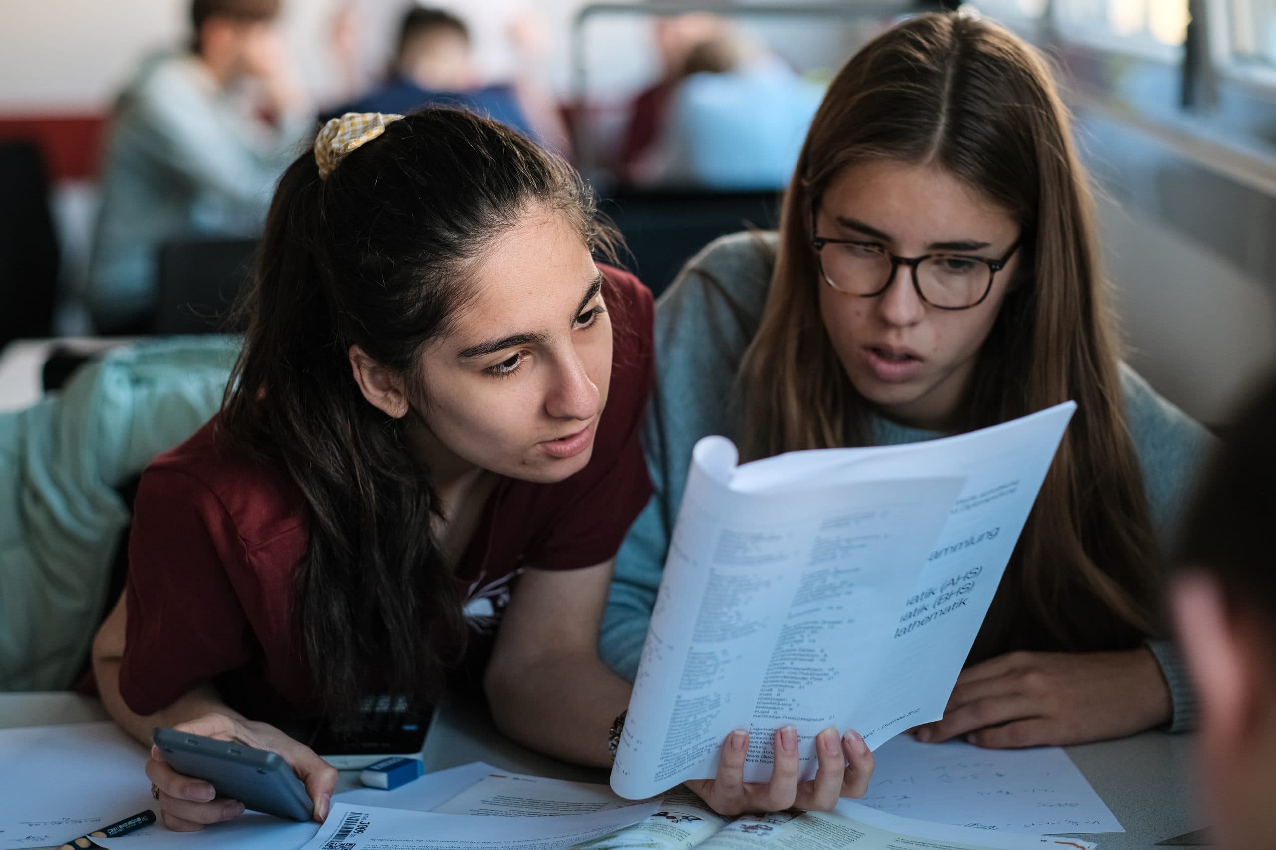 Zwei Studenten sitzen an einem Schreibtisch und studieren konzentriert ein Lehrbuch. Einer hält einen Taschenrechner in der Hand, der andere das Buch. Auf dem Tisch vor ihnen sind Papiere und Schreibmaterial ausgebreitet.