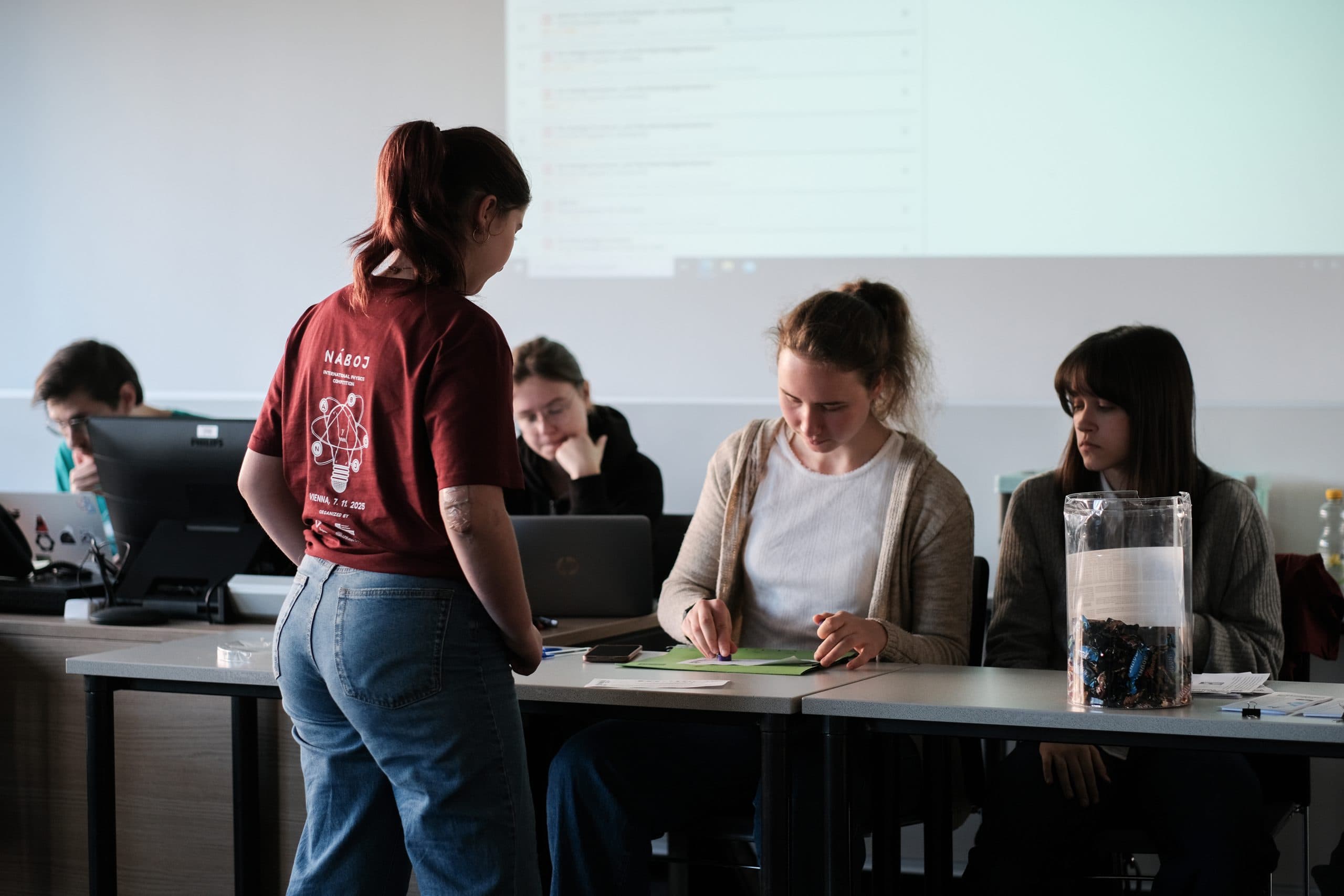 Eine junge Frau steht vor einem Tisch, an dem drei Personen sitzen, von denen eine auf einem Papier schreibt. Auf dem Tisch befinden sich Laptops, Papiere und ein durchsichtiger Behälter mit Gegenständen darin. Im Hintergrund befindet sich eine Projektionsfläche.