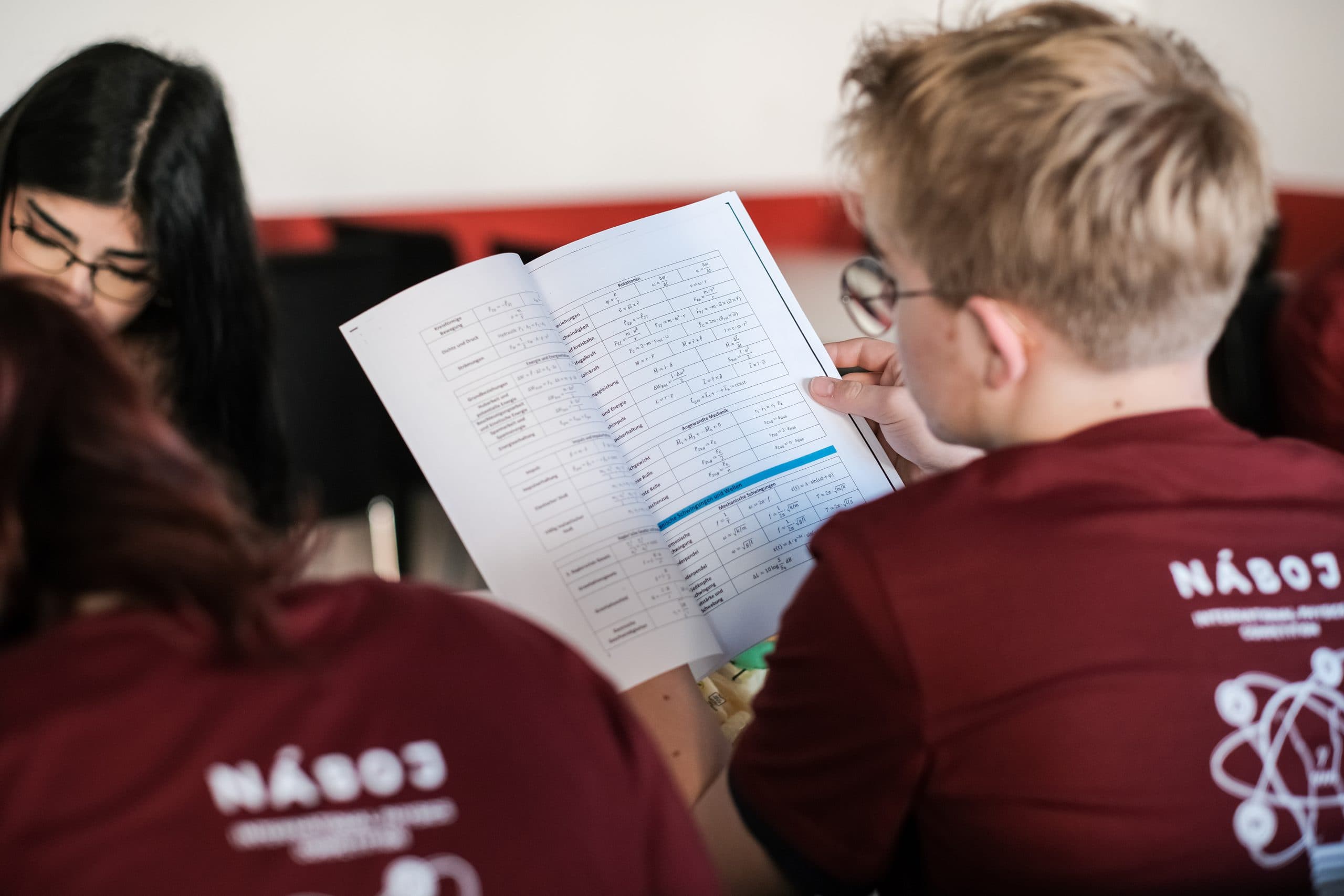 Eine Person, die eine Brille und ein rotes Hemd trägt, studiert ein Arbeitsbuch mit Diagrammen und Text, während sie mit anderen in einem Klassenzimmer sitzt. Der Fokus liegt auf den aufgeschlagenen Seiten und der Konzentration der Gruppe.
