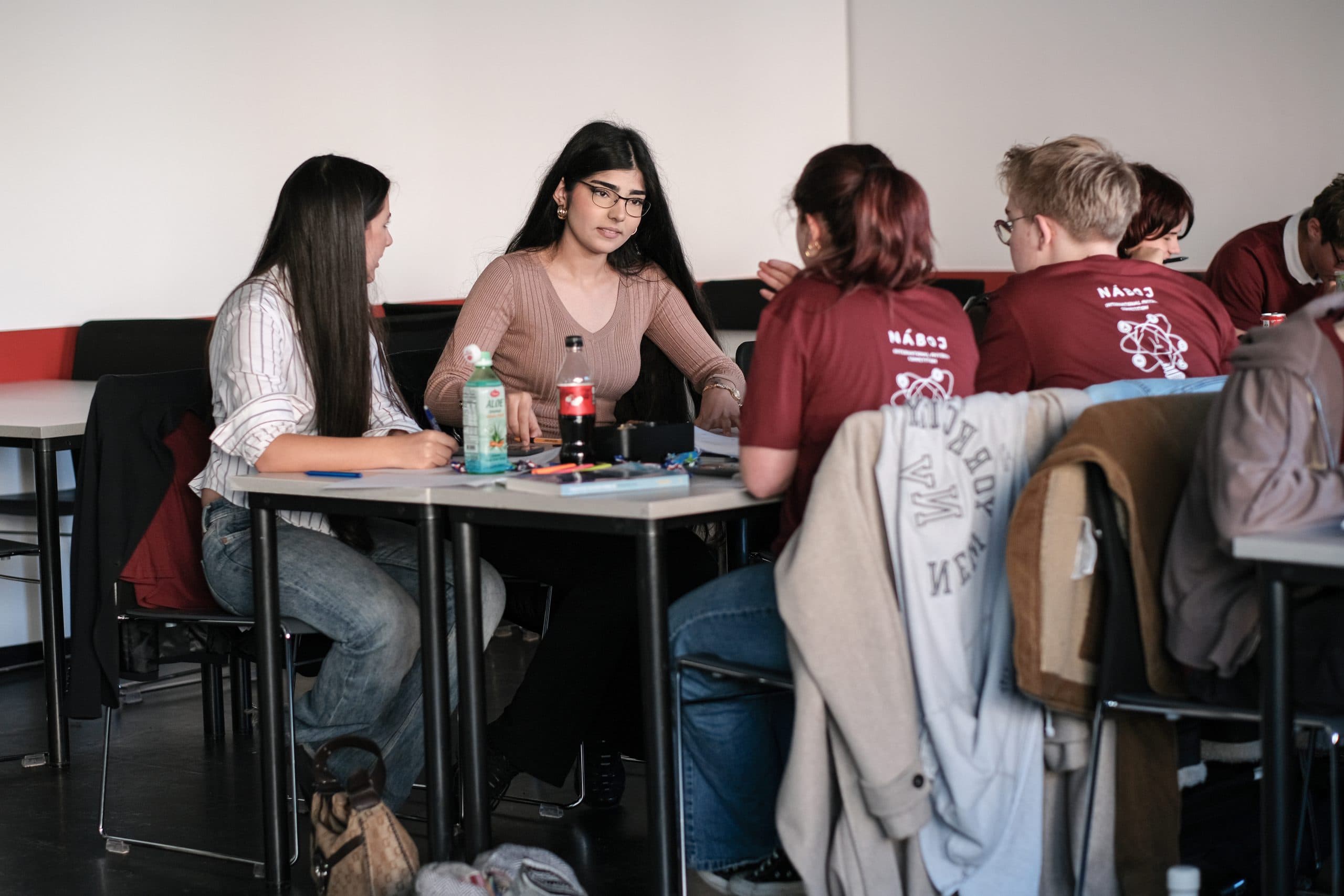 Eine Gruppe von Schülern sitzt in einem Klassenzimmer an Tischen und diskutiert. Eine Frau mit Brille spricht, während die anderen zuhören. Auf den Tischen befinden sich Getränke und Schreibwaren.