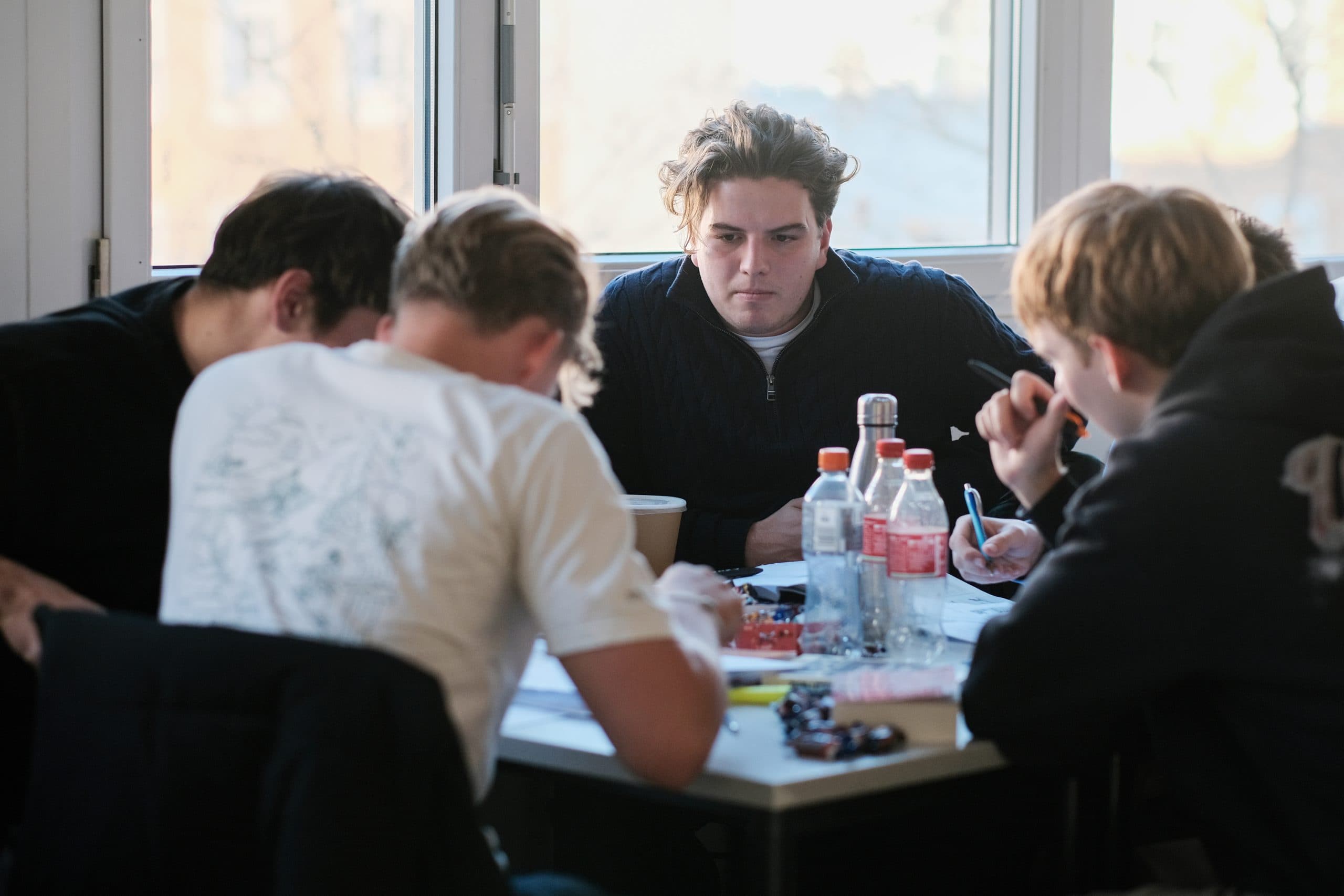 Vier junge Männer sitzen in einem sonnendurchfluteten Raum mit großen Fenstern im Hintergrund um einen Tisch, der mit Papieren, Flaschen und Snacks bedeckt ist, und konzentrieren sich auf ihre Arbeit oder ihr Studium.