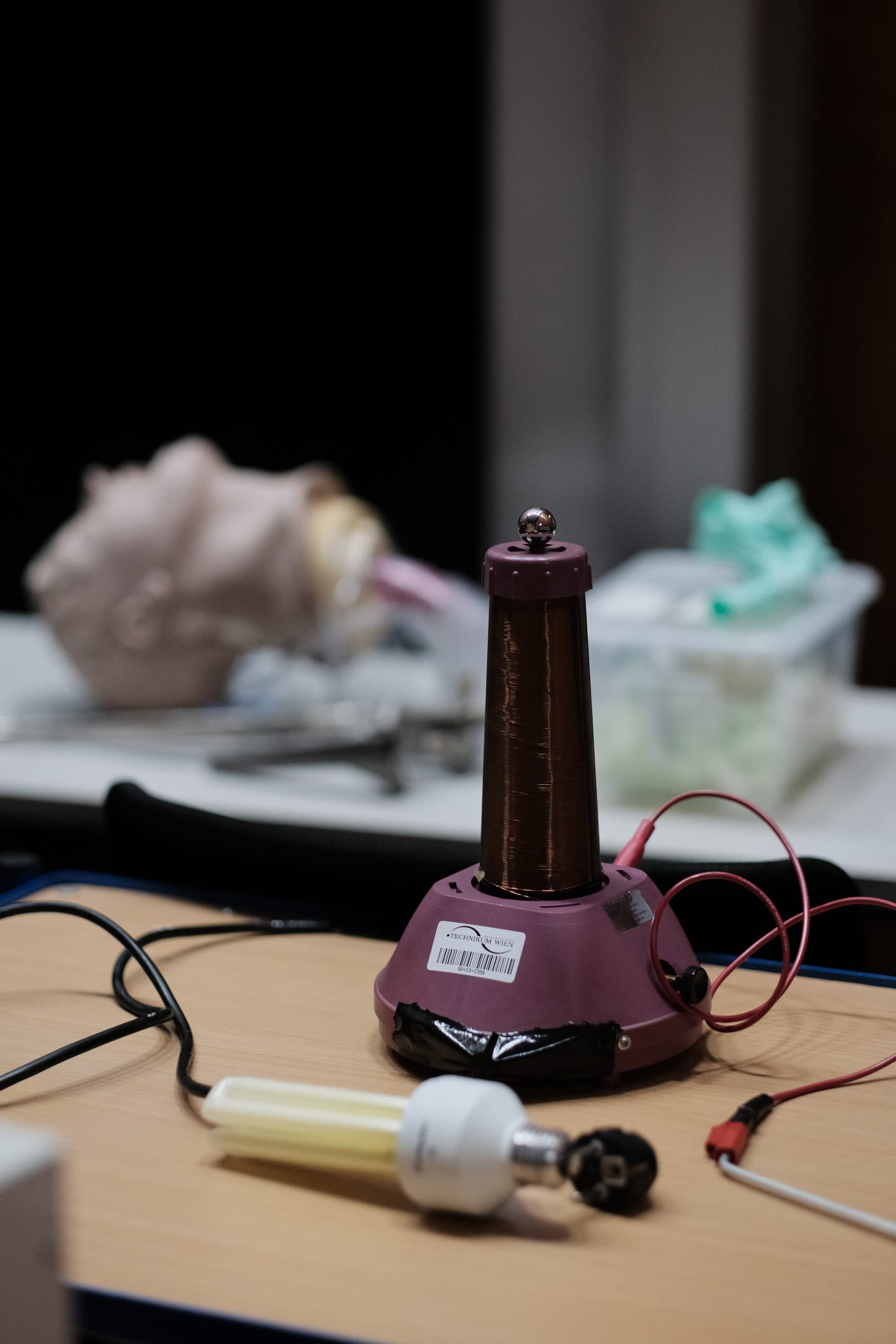Eine kleine Teslaspule mit Kupferdraht und roten Kabeln liegt auf einem Tisch, daneben eine Leuchtstoffröhre und verschiedene Drähte. Im Hintergrund sind unscharfe Objekte und Werkzeuge zu sehen.