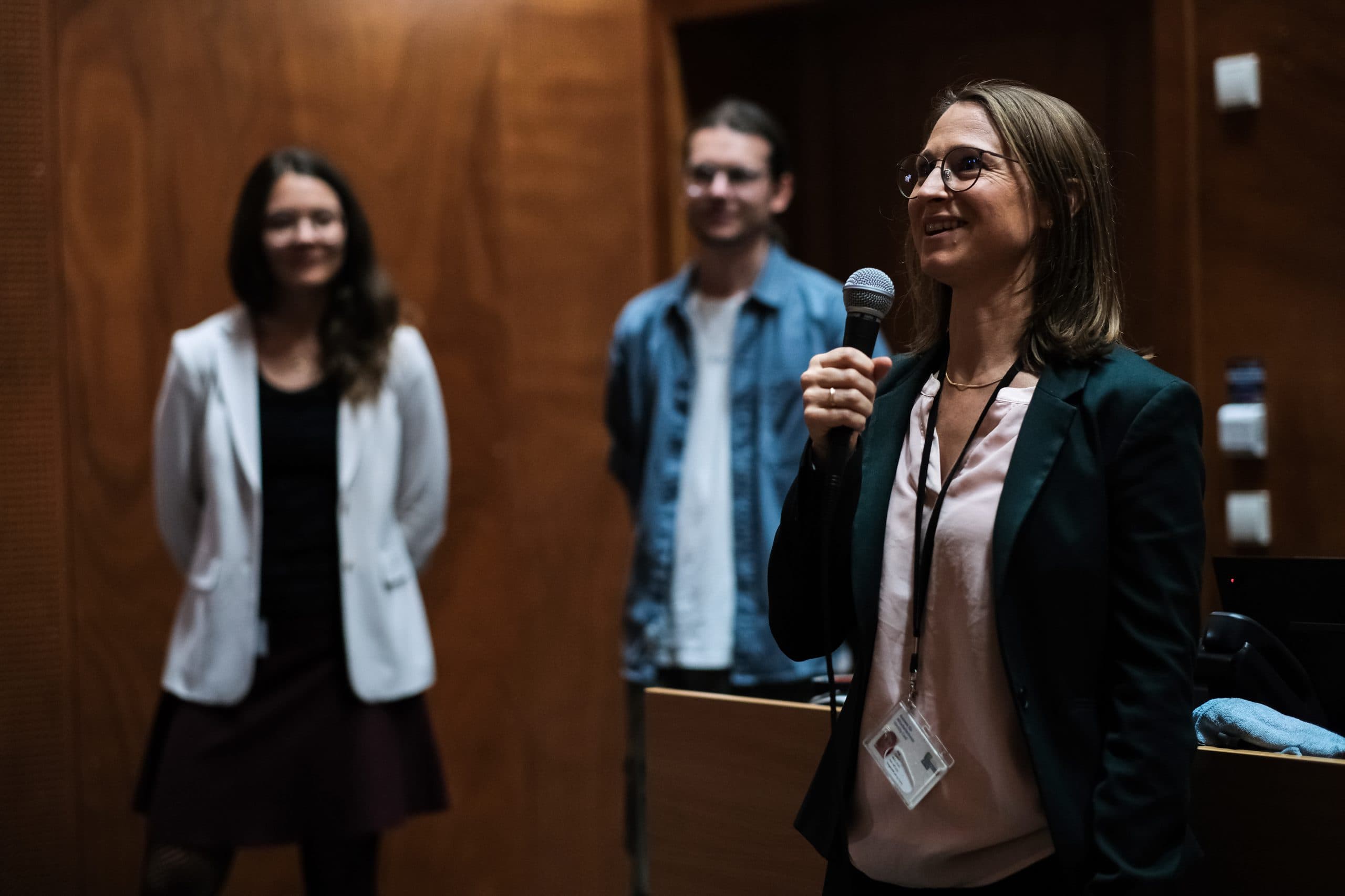 Eine Frau spricht in einem schwach beleuchteten Raum in ein Mikrofon, sie trägt eine Brille und einen Blazer. Zwei Personen stehen im Hintergrund, leicht unscharf, und hören aufmerksam zu.