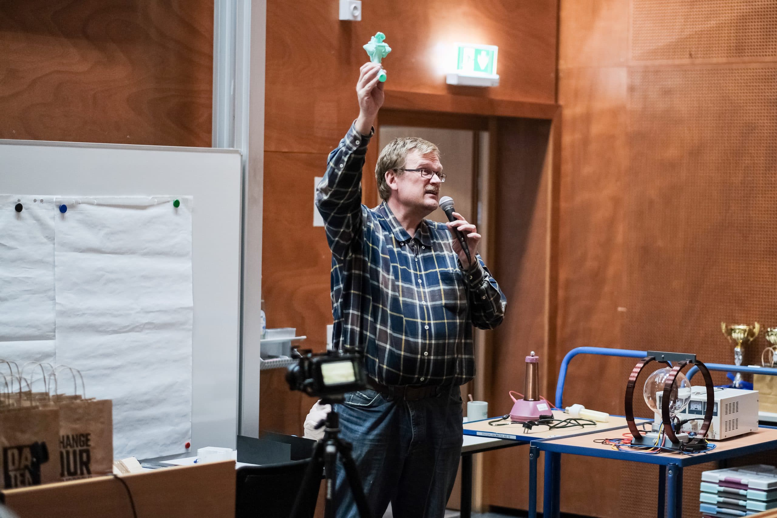 Ein Mann in einem karierten Hemd hält ein kleines grünes Objekt hoch und spricht in ein Mikrofon in einem Klassenzimmer oder Seminarraum. Um ihn herum sind ein Whiteboard, eine Kamera und verschiedene Geräte zu sehen.